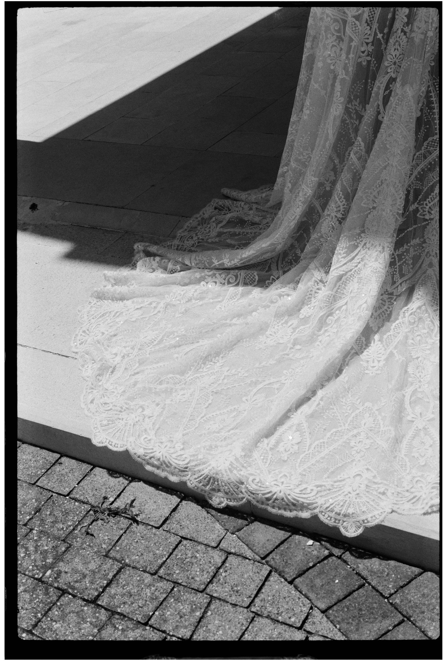 Black and white close-up of an intricate lace wedding dress train on cobblestone steps.