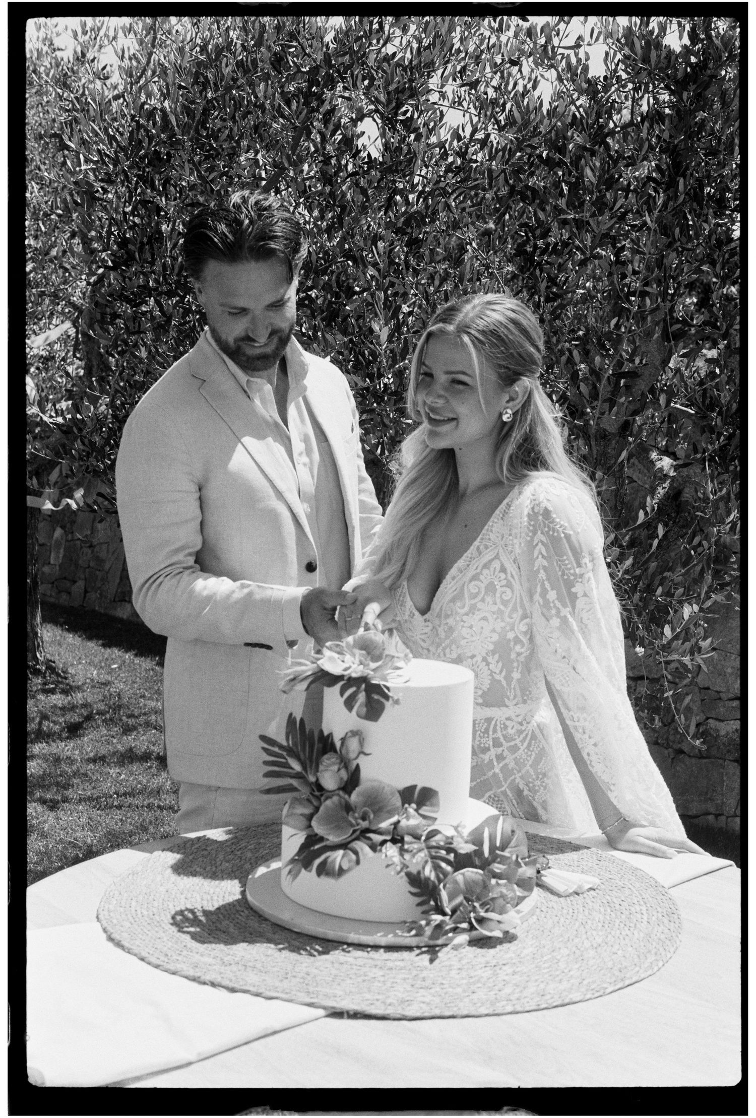 Smiling bride and groom cutting a two-tier floral wedding cake outdoors in black and white photography.