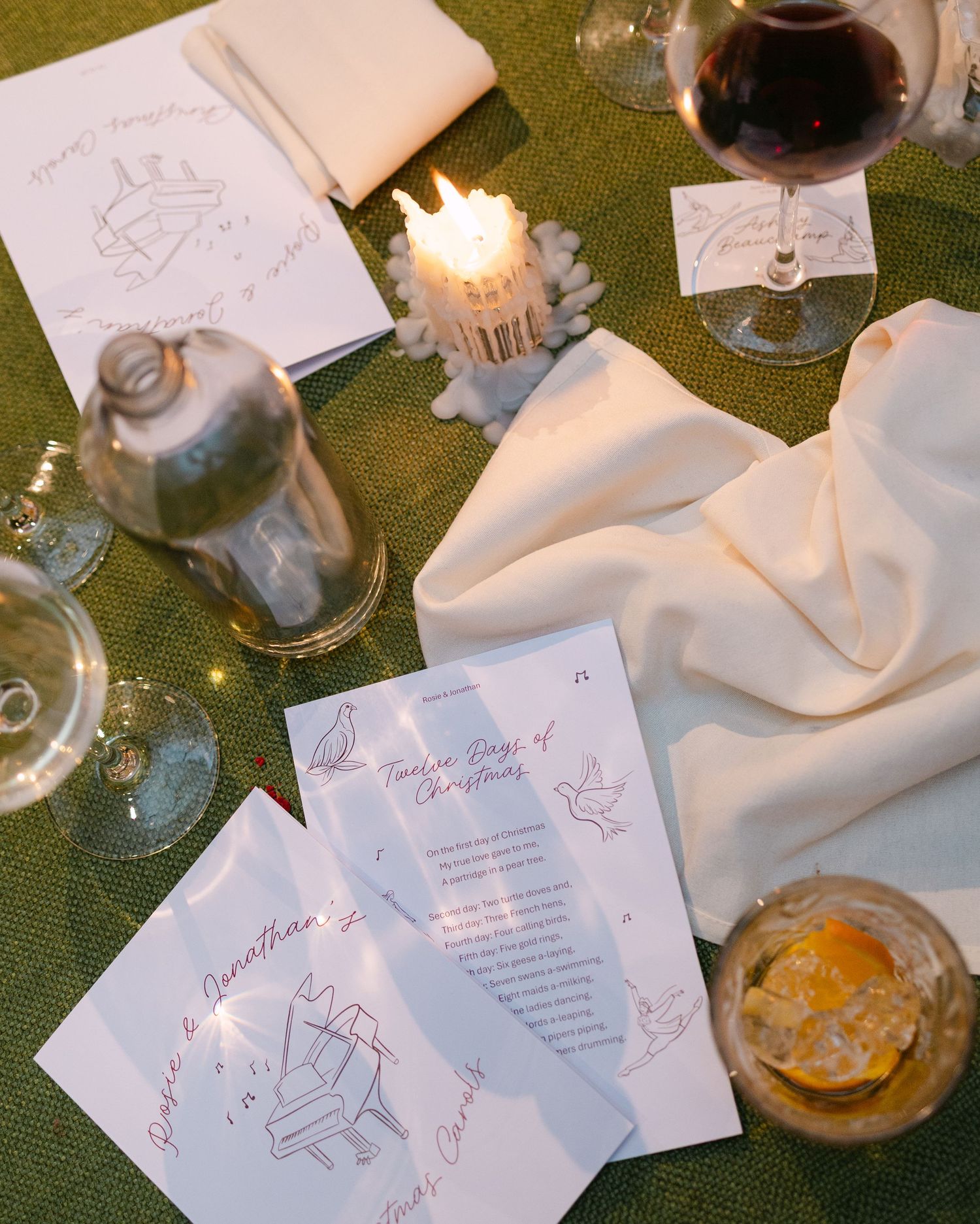 Wedding reception table with scattered menus, wine glasses, and candles on green tablecloth.