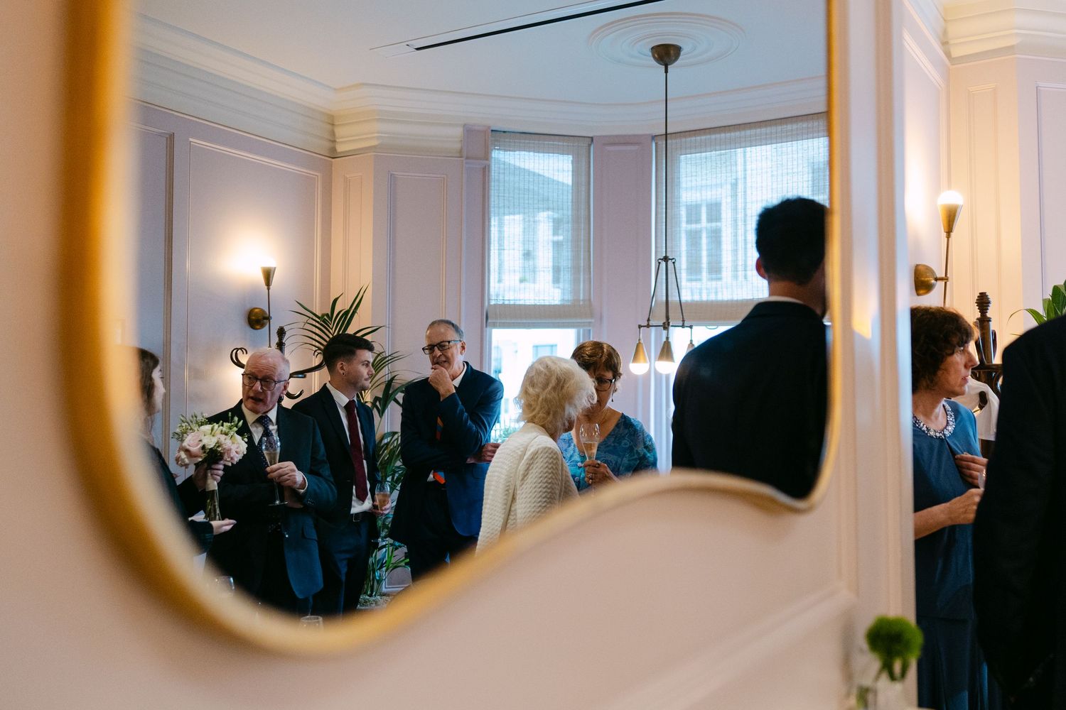 People gathered in an elegant white room with arched doorways and wall sconces for a social event at The Kensington Hotel.
