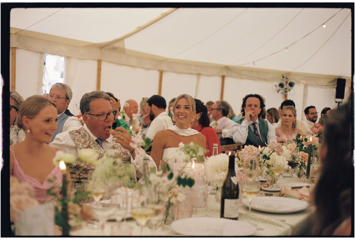 Guests laughing and celebrating at a wedding reception inside a elegant marquee tent with floral centerpieces.