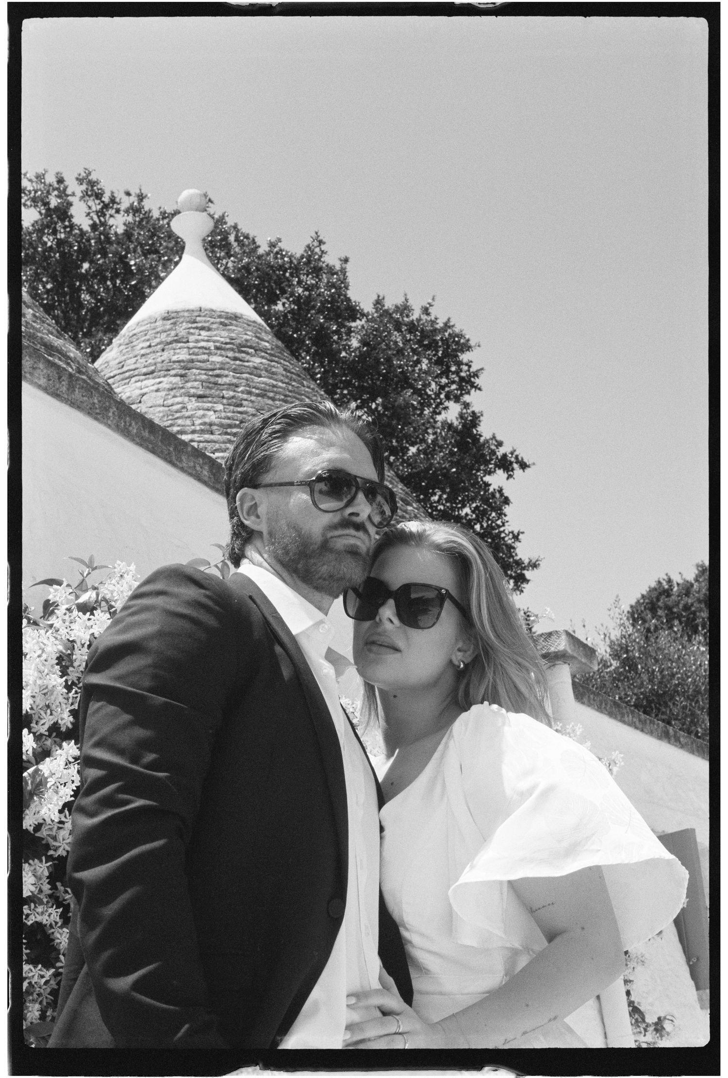 Black and white photo of an elegant couple posing outdoors near a trullo building in Italy.
