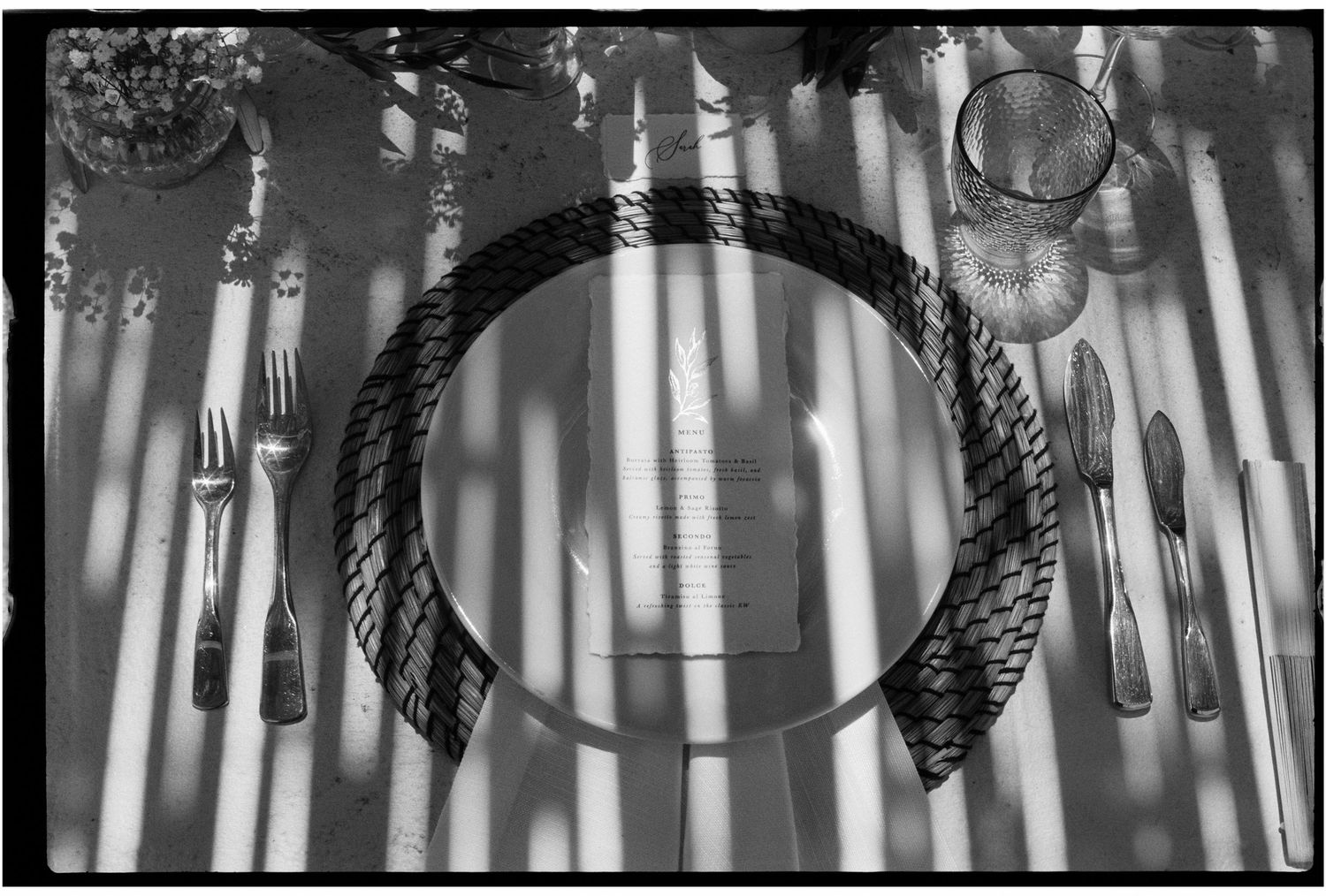 Black and white photo of an elegant table setting with rope-edged plate, silverware, and glassware casting striped shadows.