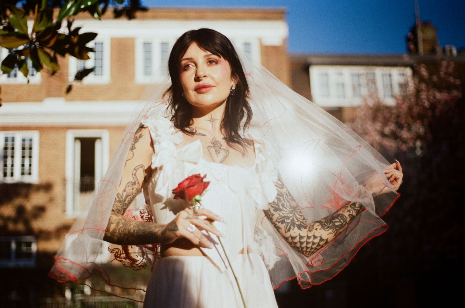 Tattooed bride in white dress and veil holds red rose outdoors on sunny day.