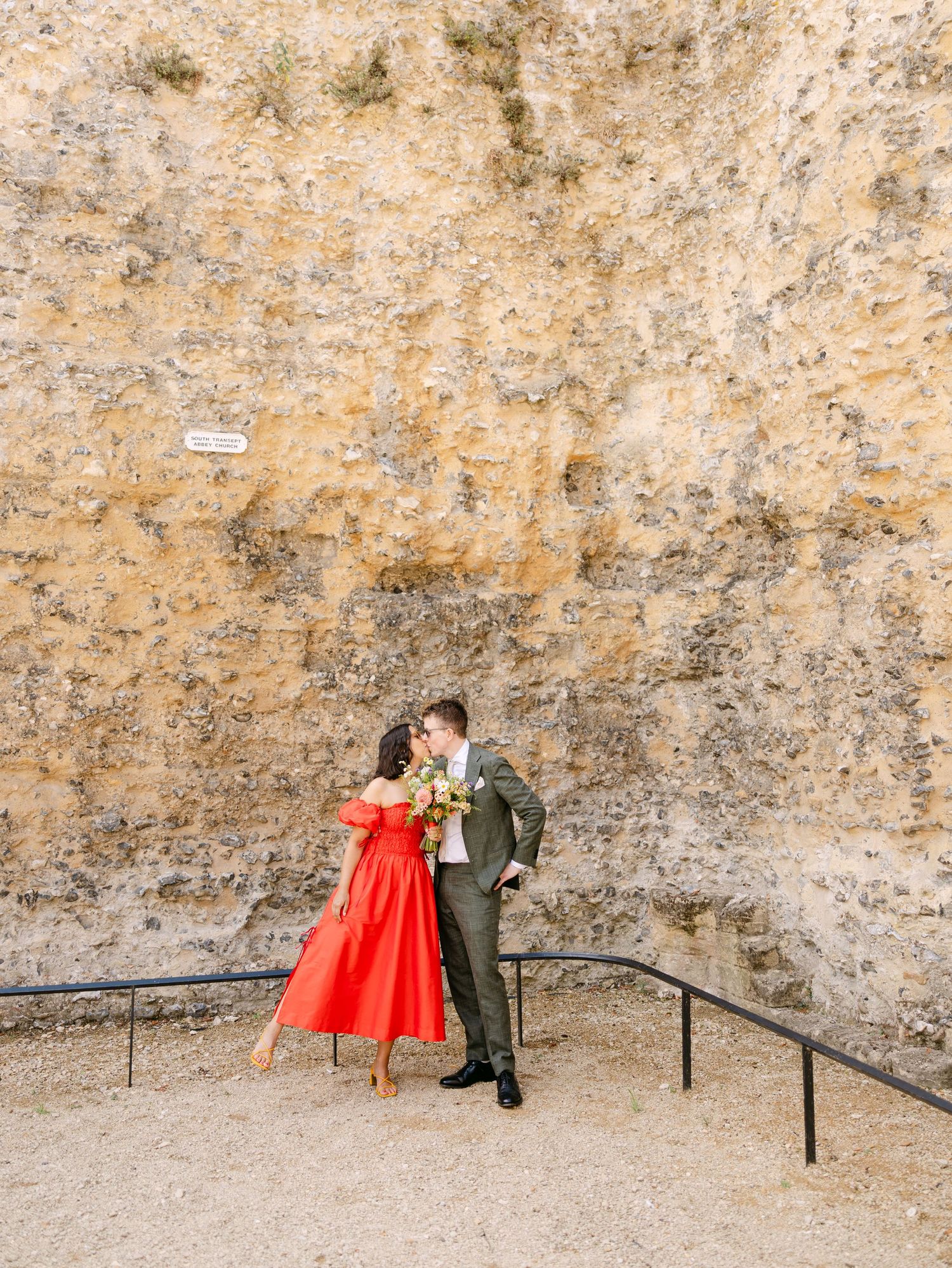 Couple kissing in red dress and olive suit against a textured stone wall, holding a colorful bouquet outdoors.