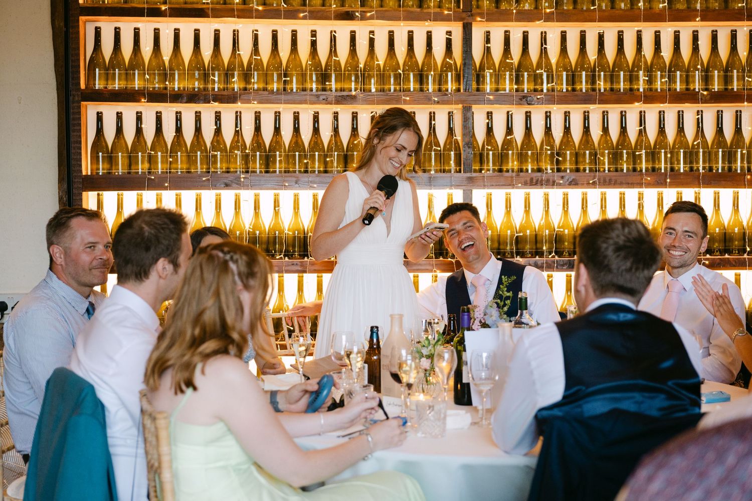 Bride giving a speech at wedding reception with wine bottle wall backdrop, guests laughing at table.