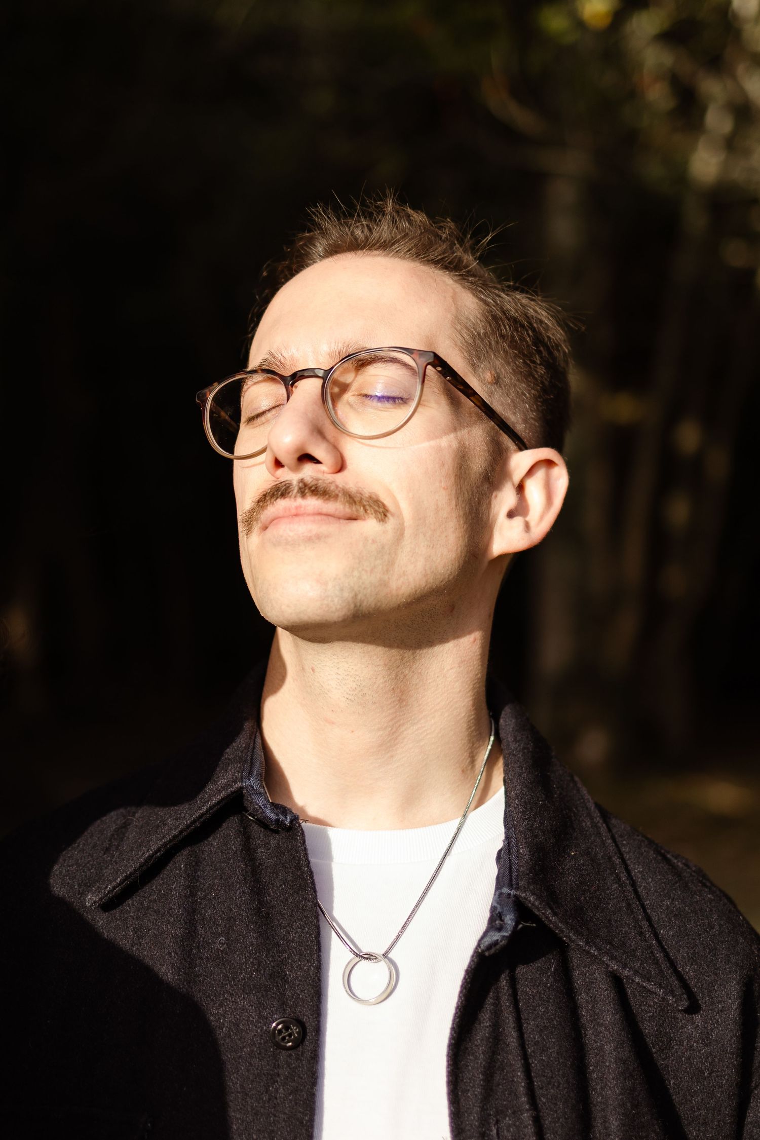 Person in glasses and dark coat smiling with eyes closed in natural sunlight.