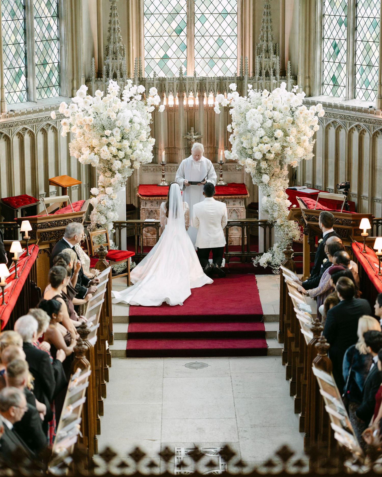 Wedding ceremony in progress inside historic church with stained glass windows and white floral arrangements at Ashridge House.