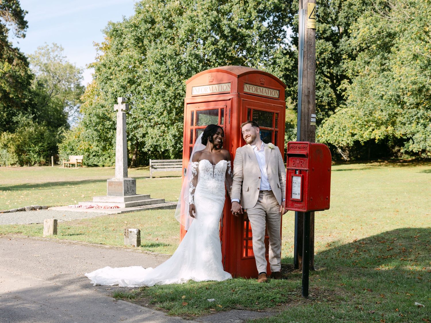A couple in wedding attire poses for photos near a classic red British phone booth and postbox in a park setting.