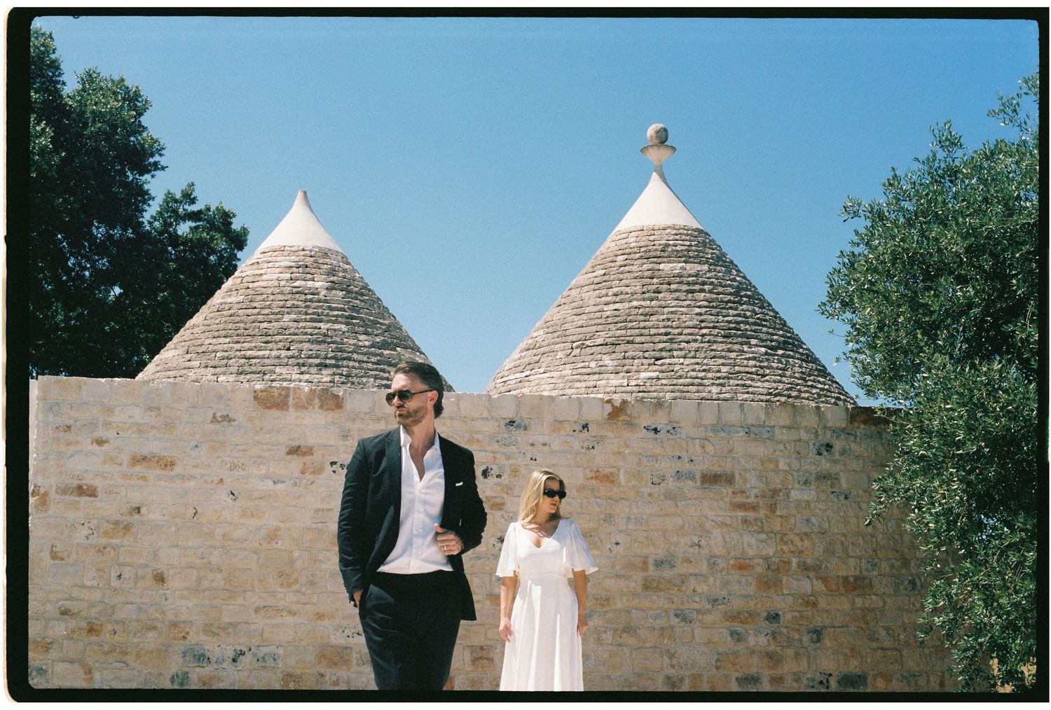 Film photo of a couple walking hand in hand past traditional trulli stone buildings under a blue sky.