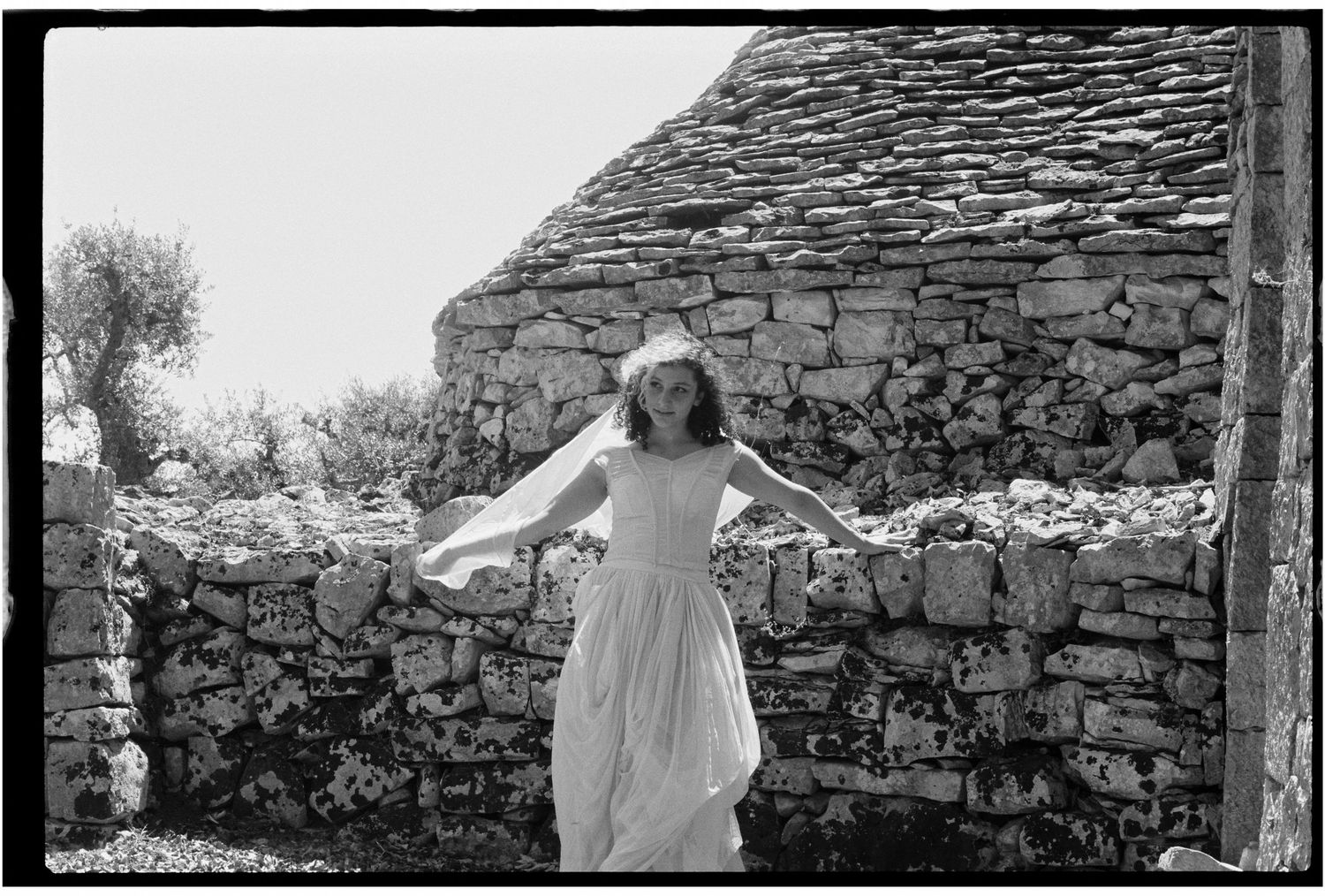 Woman in white dress poses gracefully against rustic stone wall ruins in black and white photography.