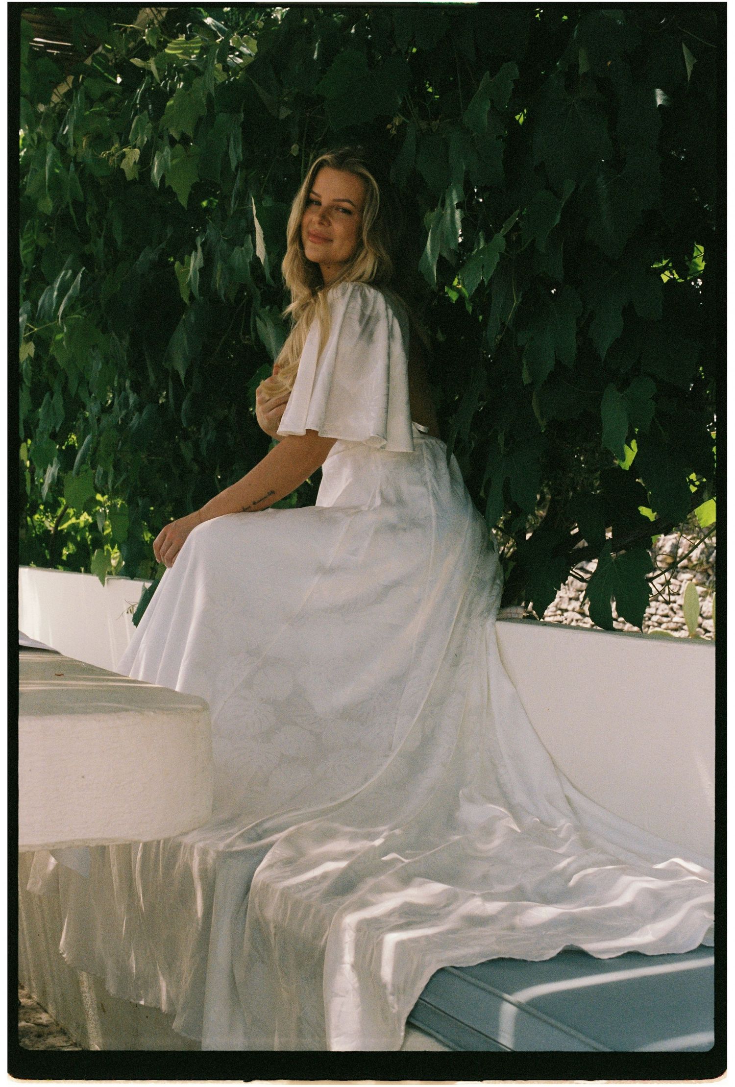Bride in a flowing white gown sitting on a stone wall surrounded by green foliage.