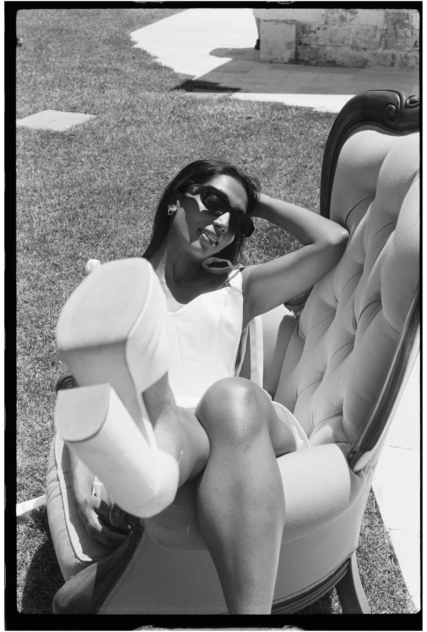 Black and white photo of a woman in white outfit and sunglasses relaxing in an elegant chair outdoors in the sun.