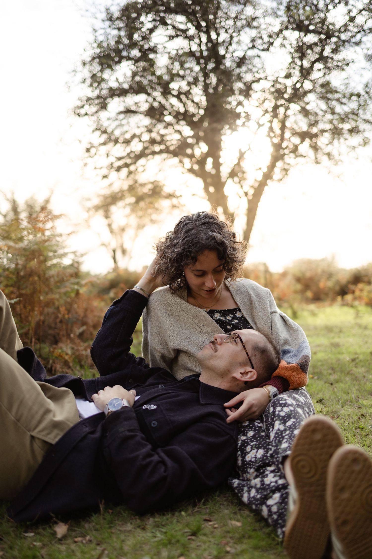 A couple relaxes together on a grassy hill during sunset with trees silhouetted in the background.