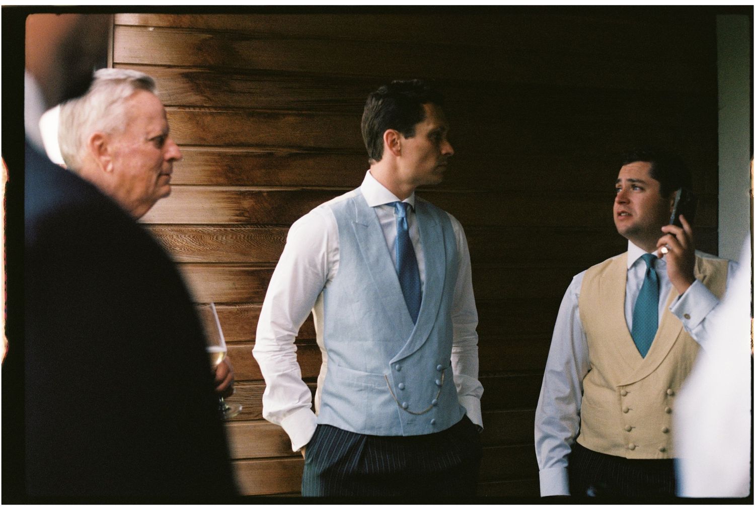 Two groomsmen in vests and ties stand near a wooden wall, one talking on phone, at a wedding venue.