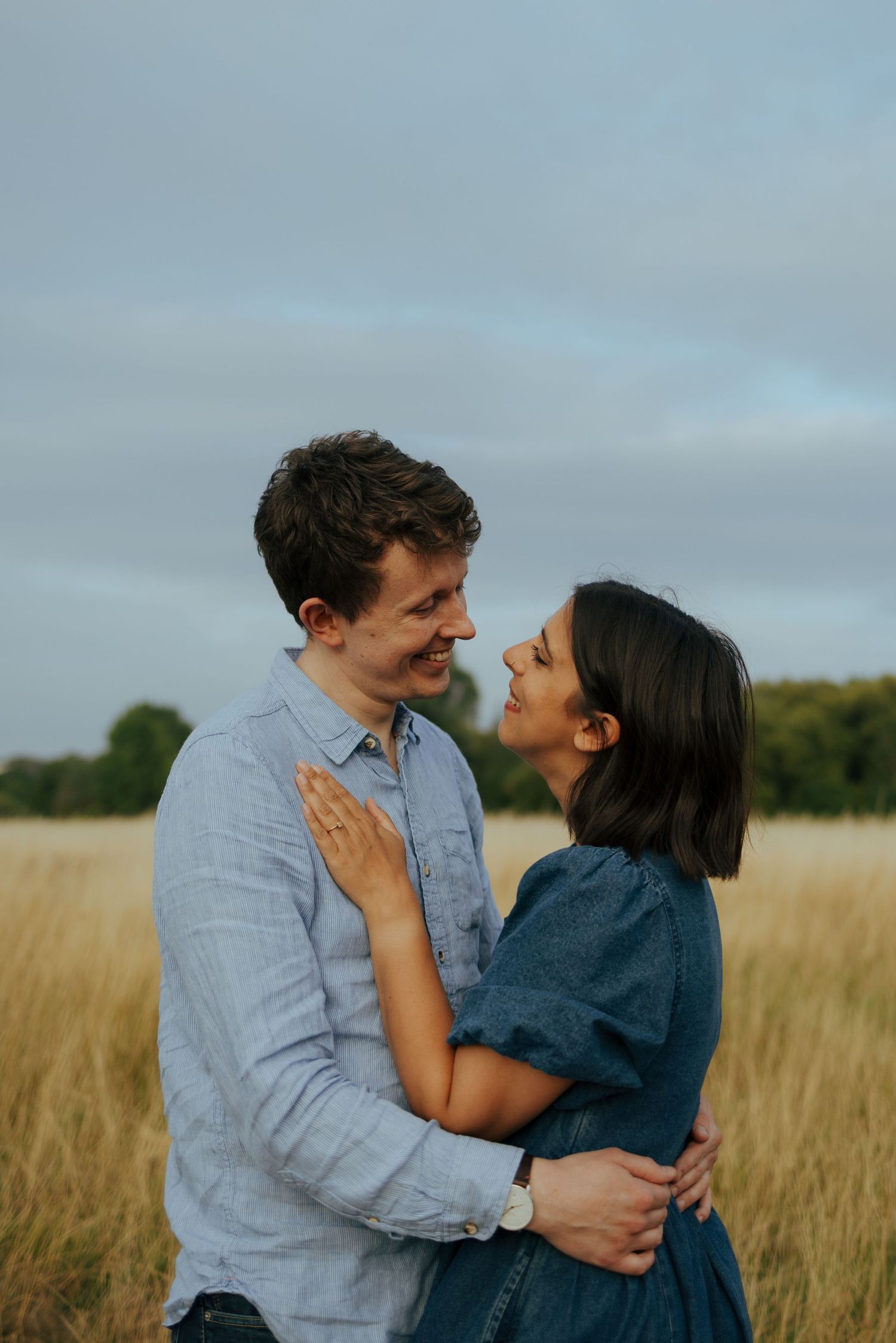 A couple embraces and laughs together in a field of tall grass at sunset.