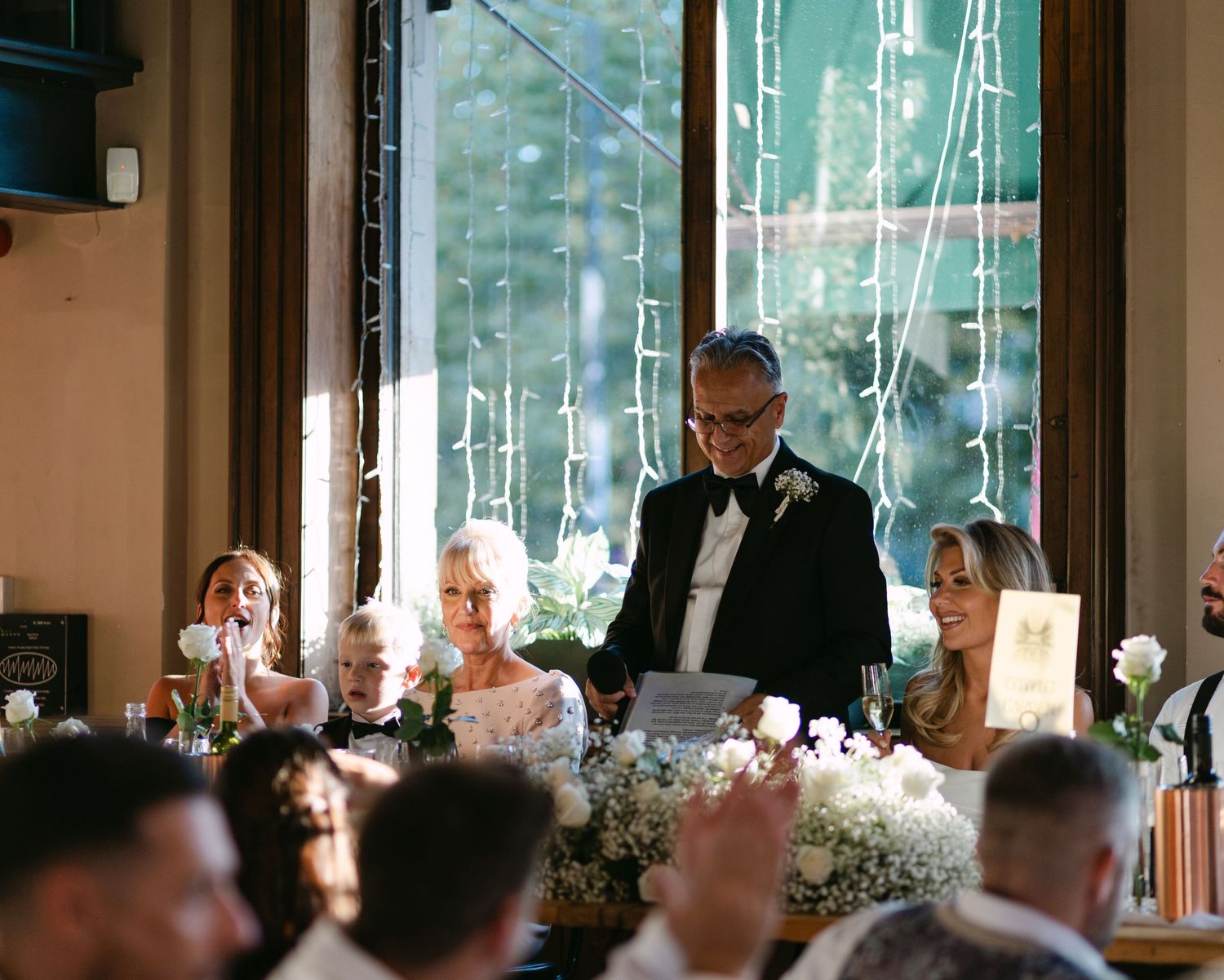 A speaker delivers a toast during an intimate wedding reception with floral centerpieces.