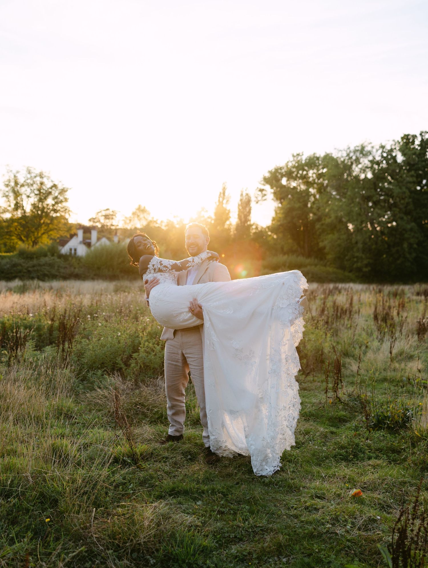 A couple shares joyful laughter in a sunlit field wearing wedding attire.
