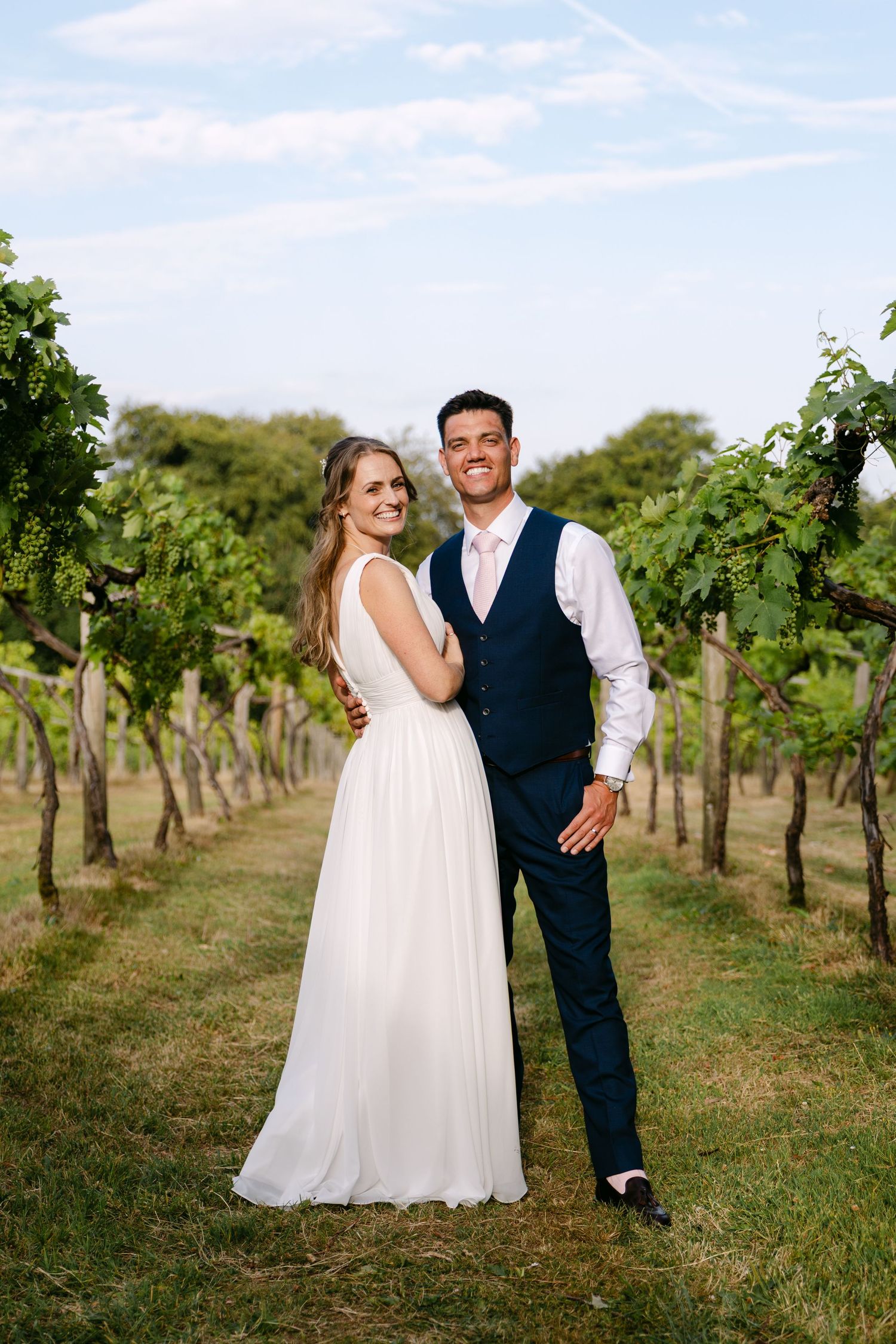 Smiling bride and groom pose together in a lush vineyard on their wedding day.