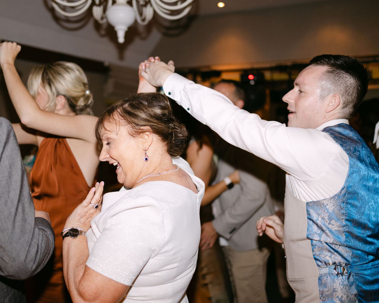Wedding party dancing with raised arms in celebration at indoor reception.