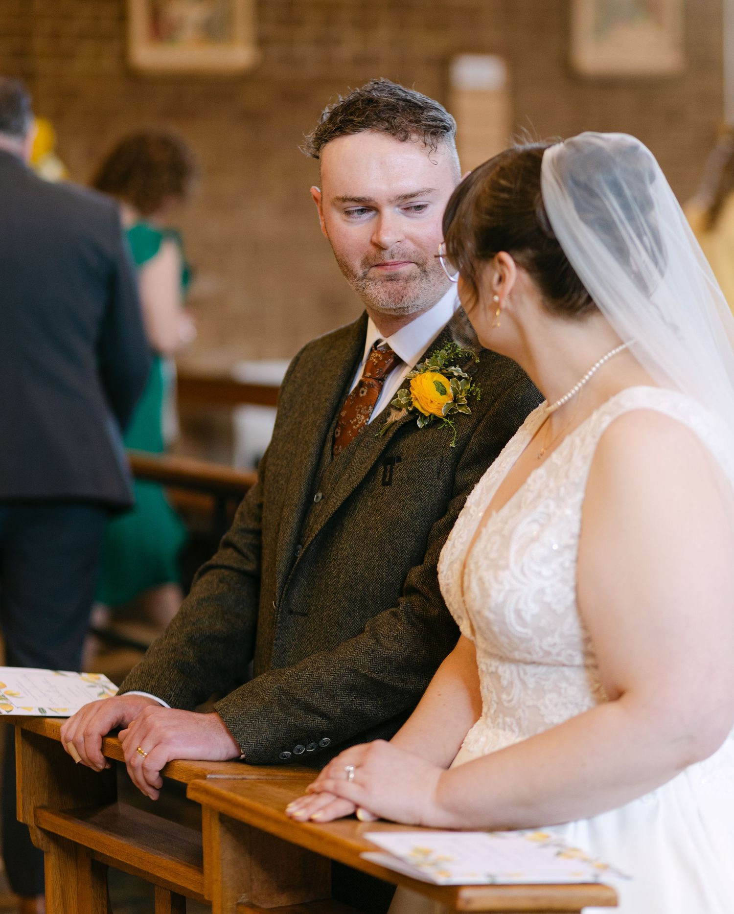 Bride and groom sit together in church pew during wedding ceremony, groom wearing green tweed suit with yellow floral buttonhole.