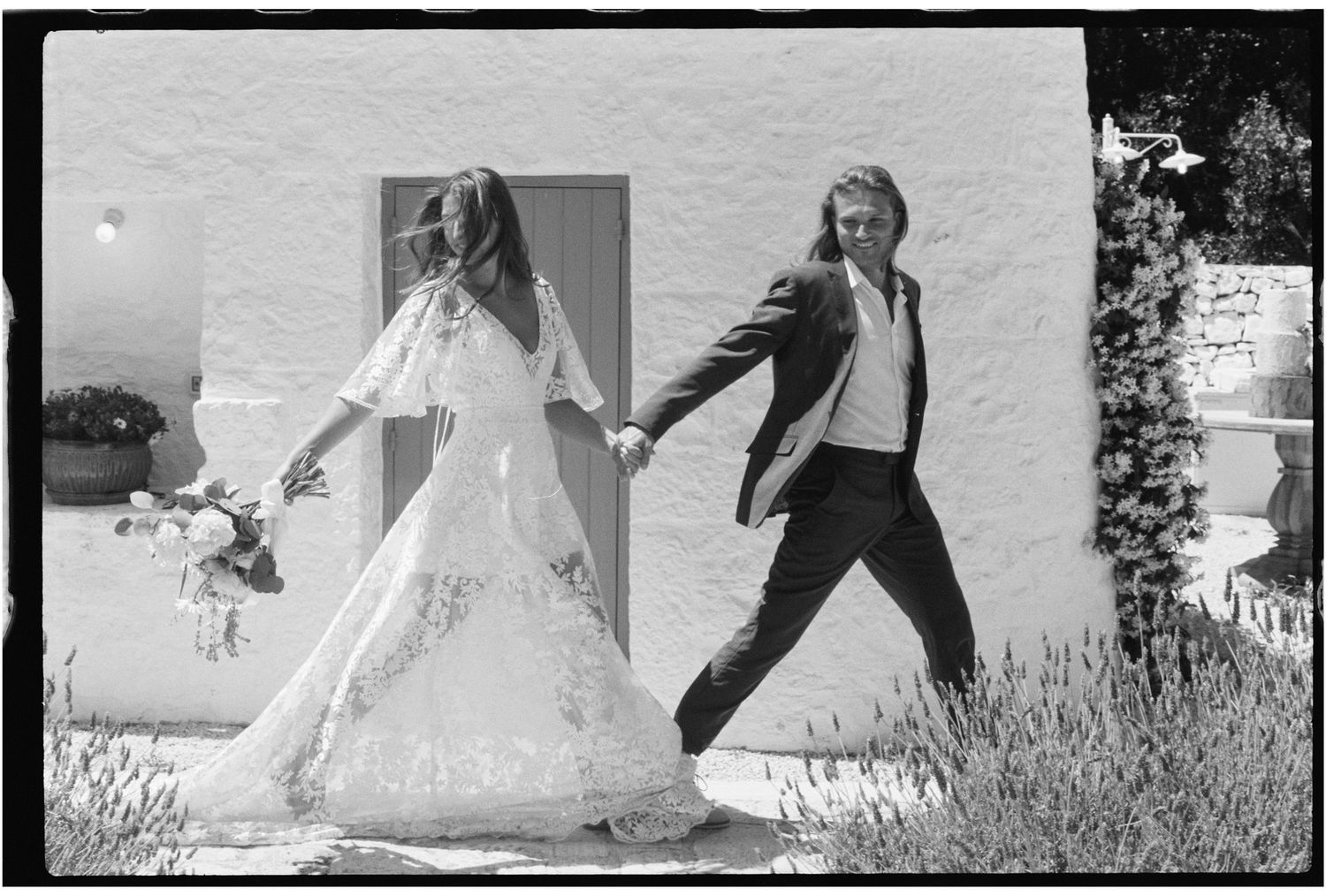 Black and white photo of a bride in lace gown and groom in suit holding hands outdoors at a wedding.