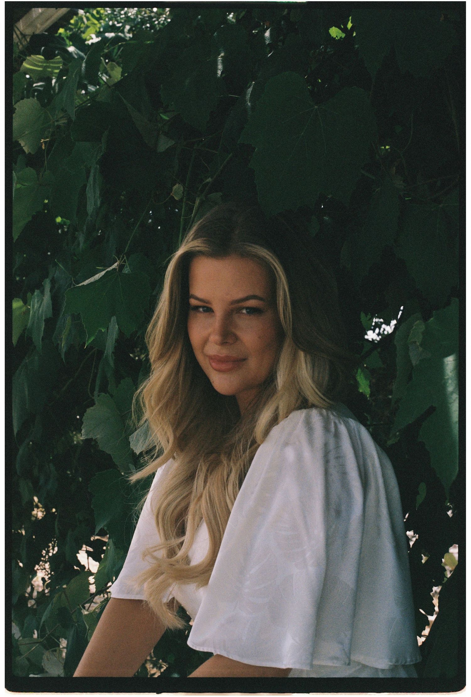 Film portrait of a smiling blonde woman in a white top surrounded by lush green leaves outdoors.