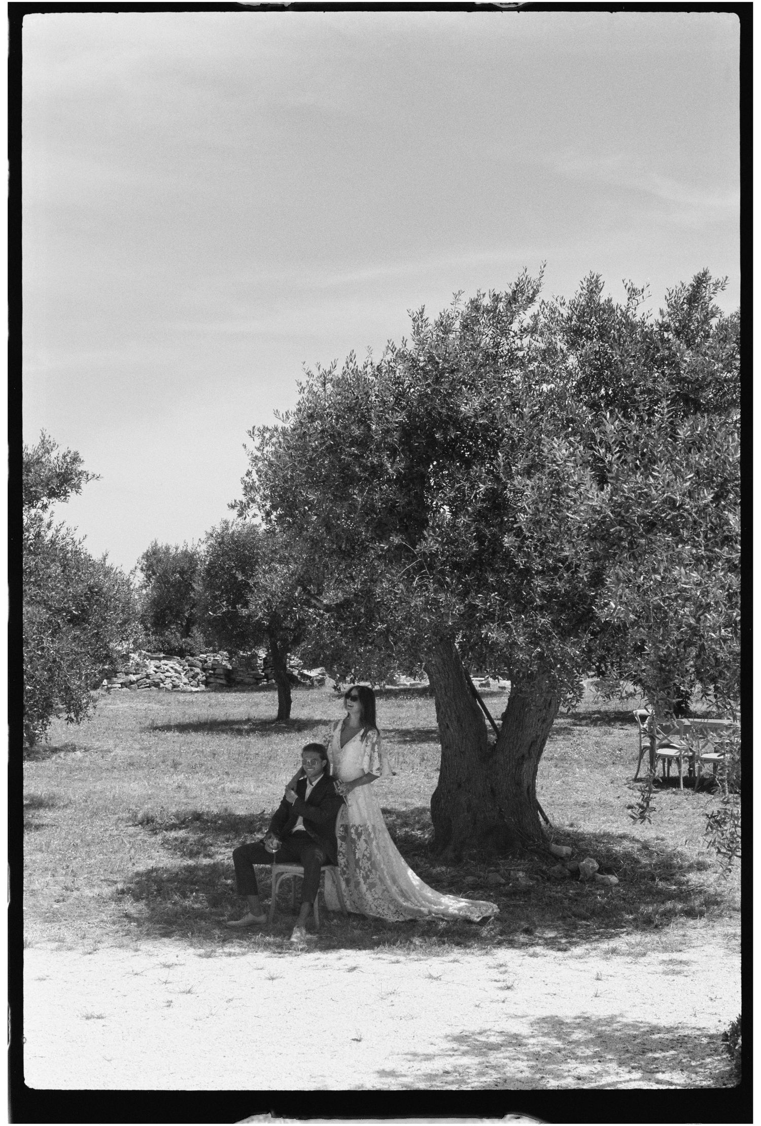 Bride and groom sitting together under an olive tree in a sunlit Mediterranean grove, black and white wedding photo.