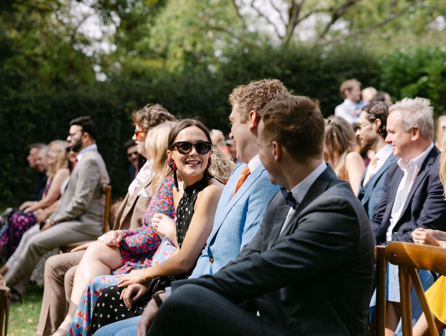 Wedding guests seated outdoors on chairs enjoying a sunny ceremony.