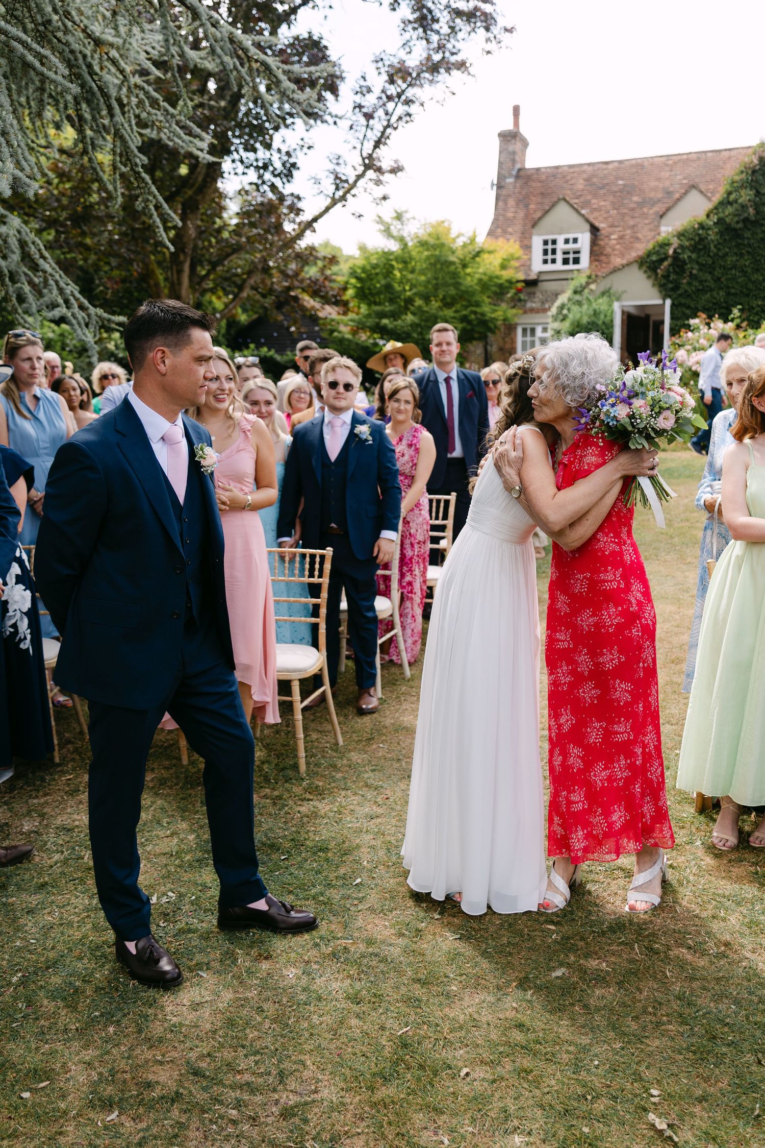 Bride embracing a guest in red dress at outdoor garden wedding ceremony while groom watches in navy suit.