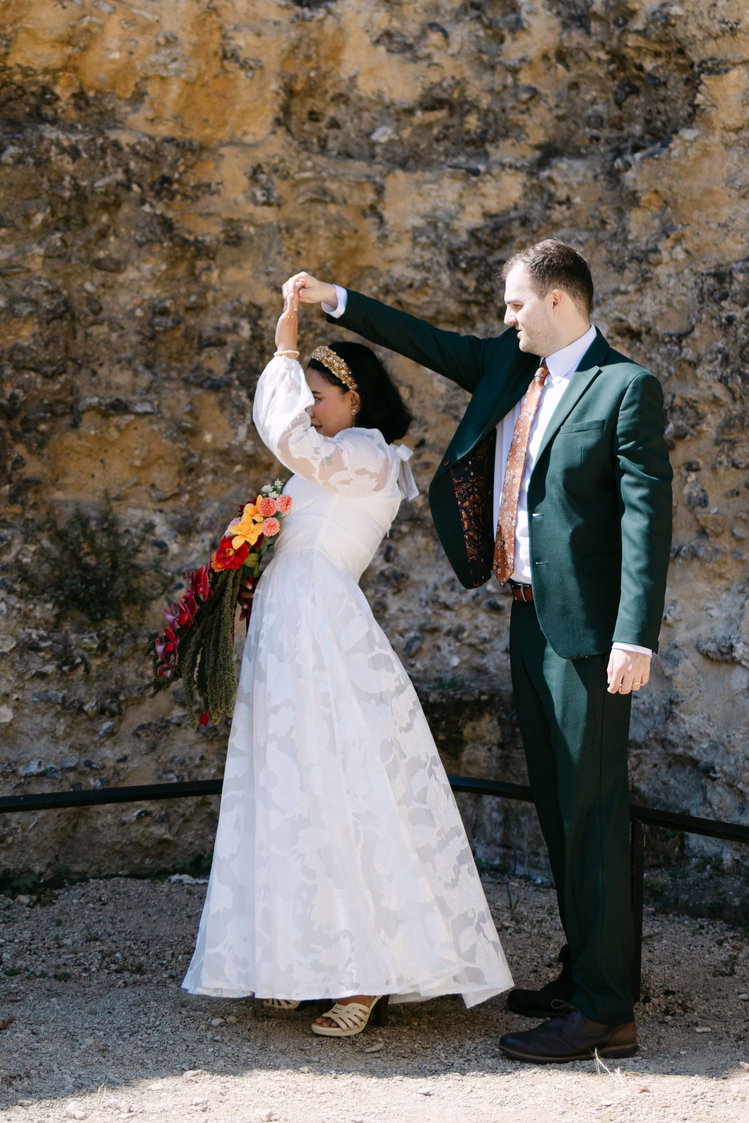 A bride in white gown and groom in green suit dance outdoors near a stone wall, holding colorful bridal bouquet.