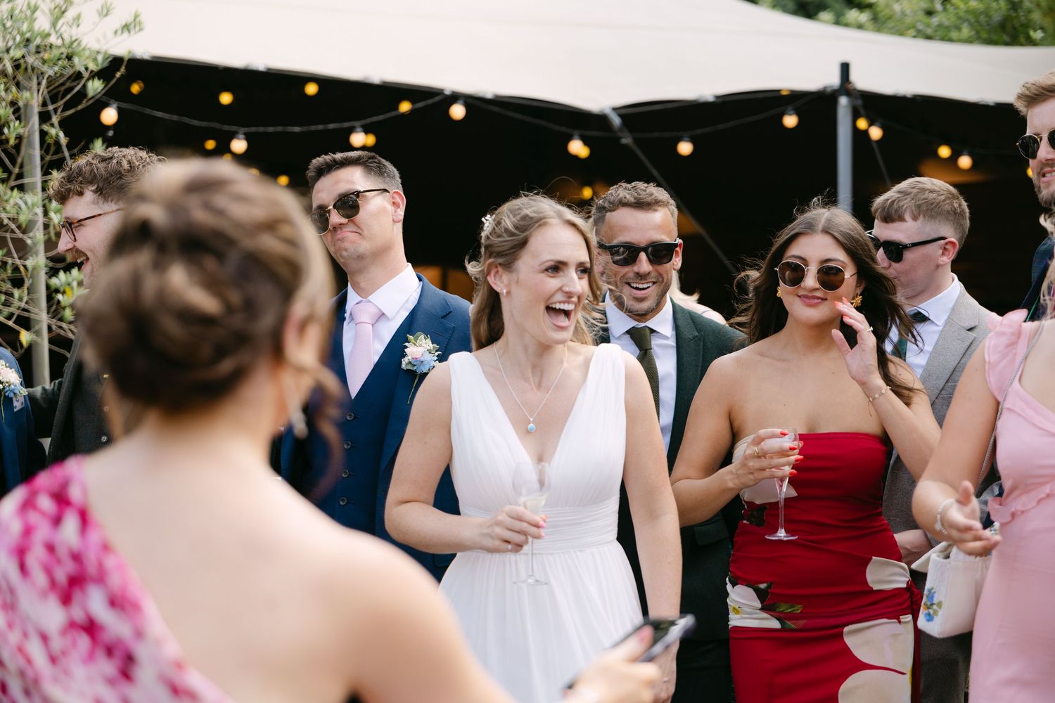 Bride in white dress laughing with wedding guests outdoors, some wearing sunglasses, at a sunny garden reception.