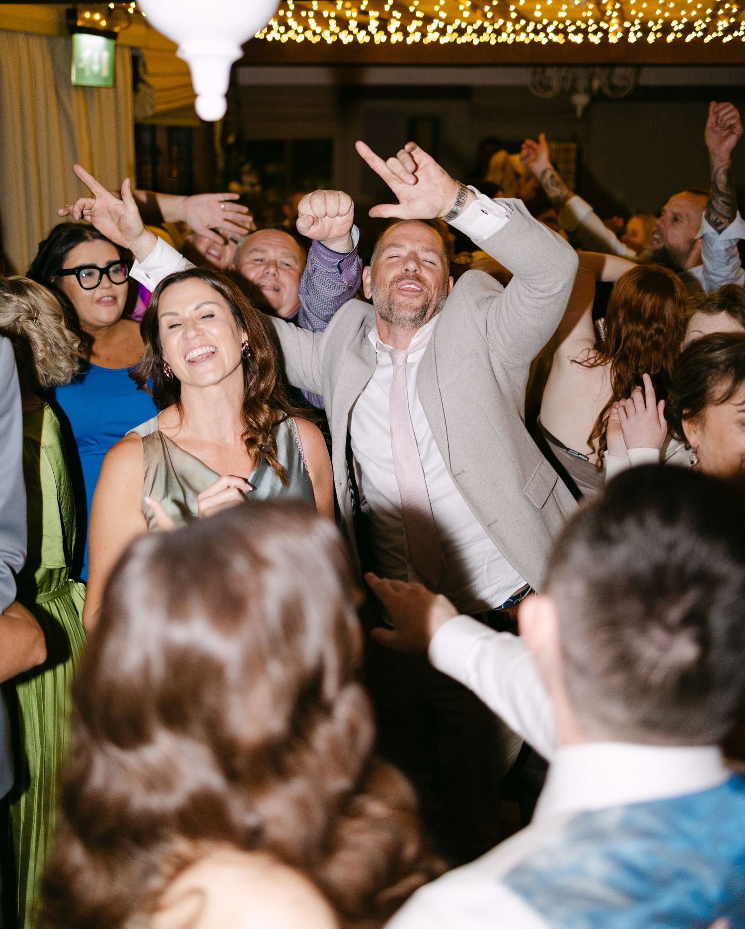 Energetic crowd dancing and celebrating at a wedding reception under string lights.