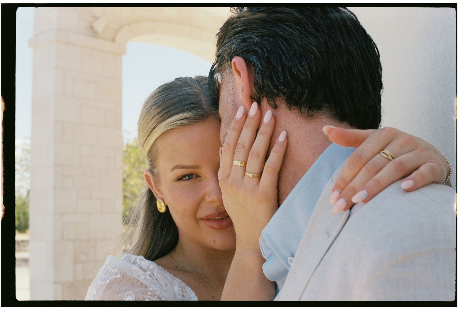 Smiling blonde bride embracing groom near stone columns, showing off her gold wedding ring.