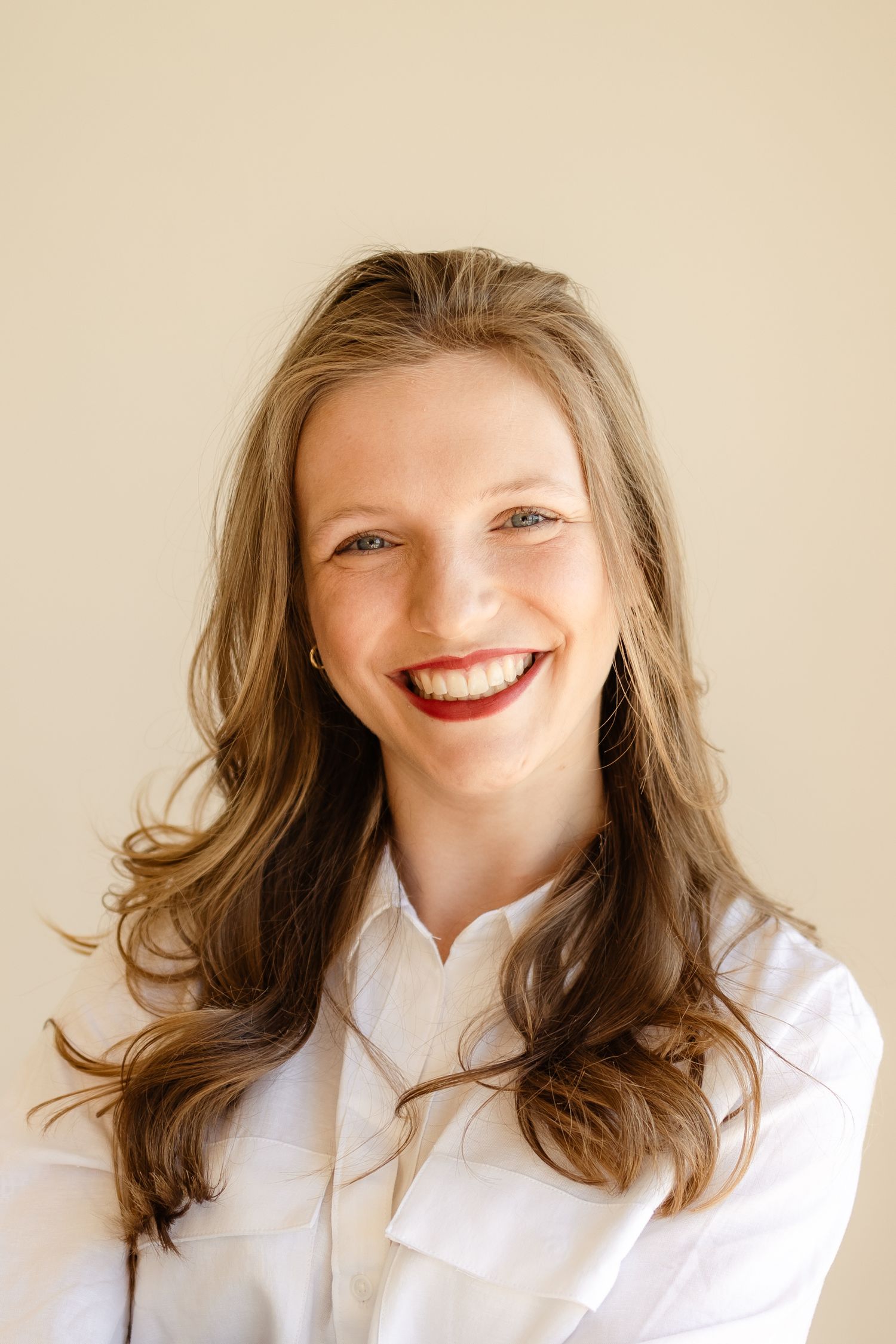 Smiling young woman with long brown hair wearing a white shirt against a beige background.