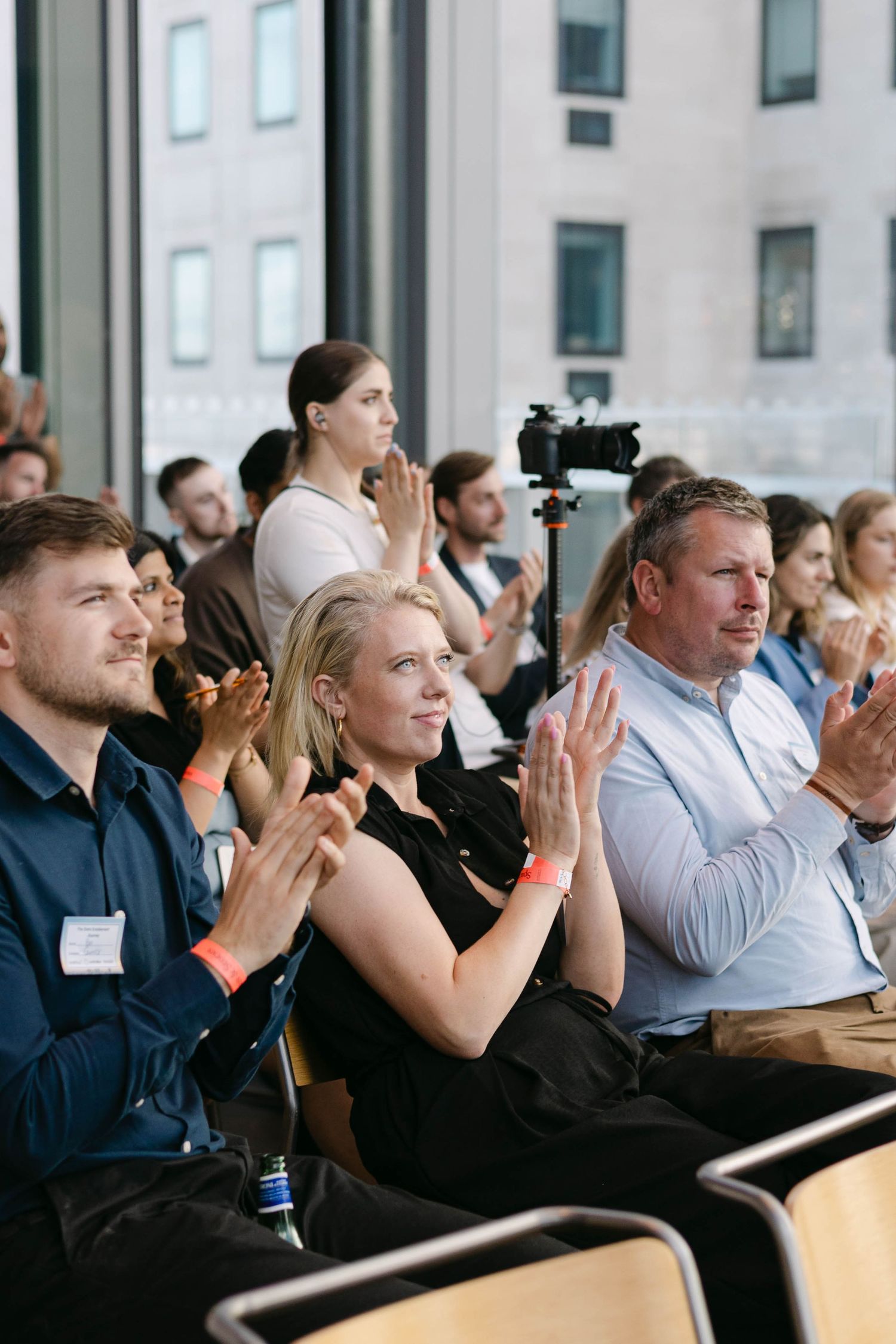 Audience members with orange wristbands applaud at a professional conference event in a modern building.