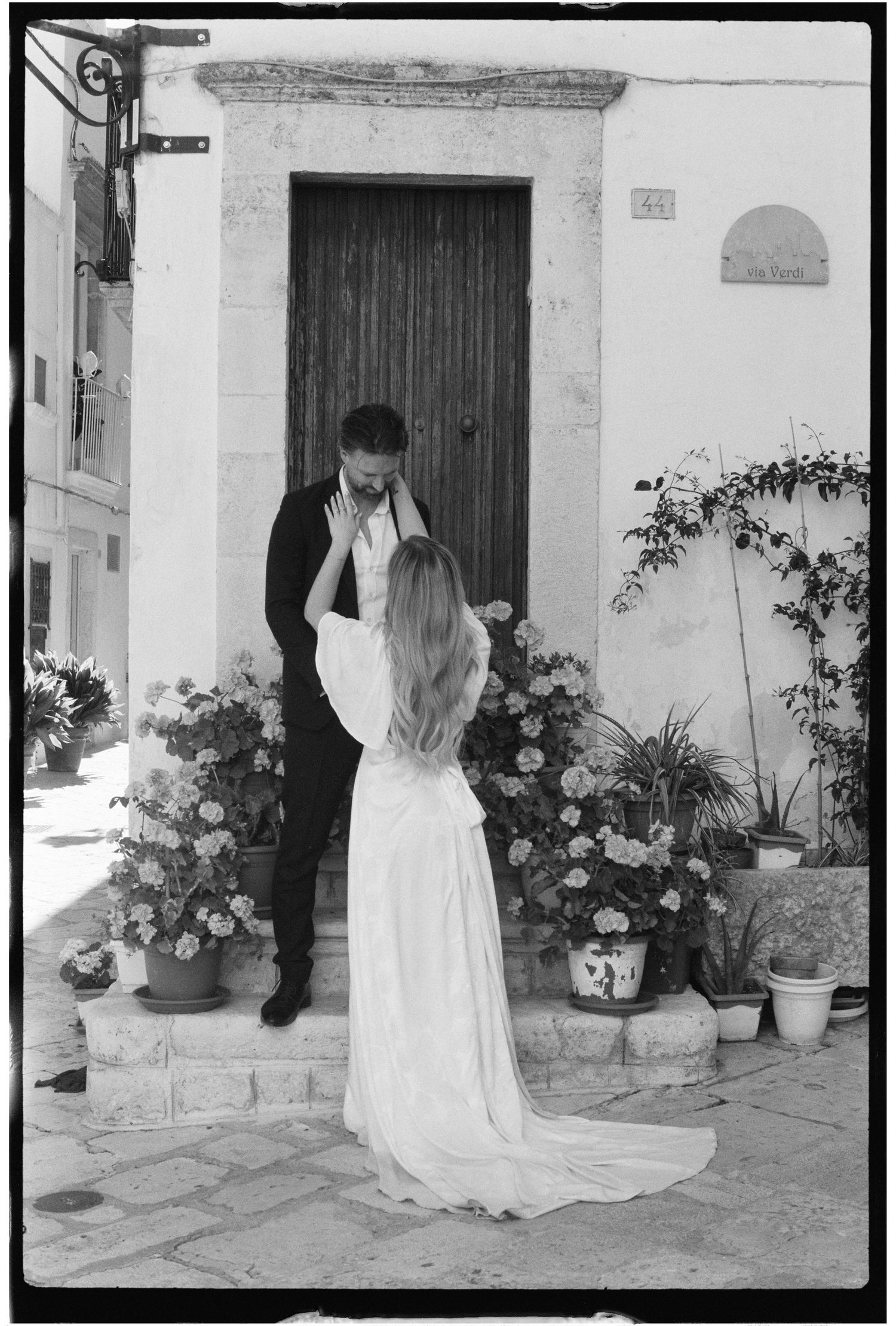 Black and white photo of a bride and groom sharing a tender moment outside a rustic European doorway with flowers.