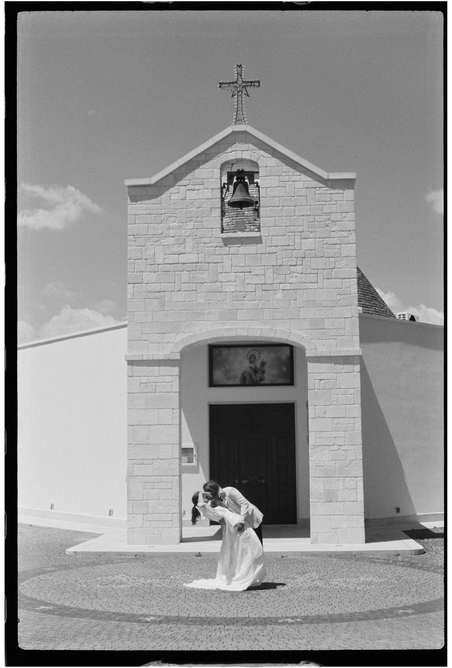 Couple sharing a kiss in front of a small stone chapel with a bell and cross in black and white.