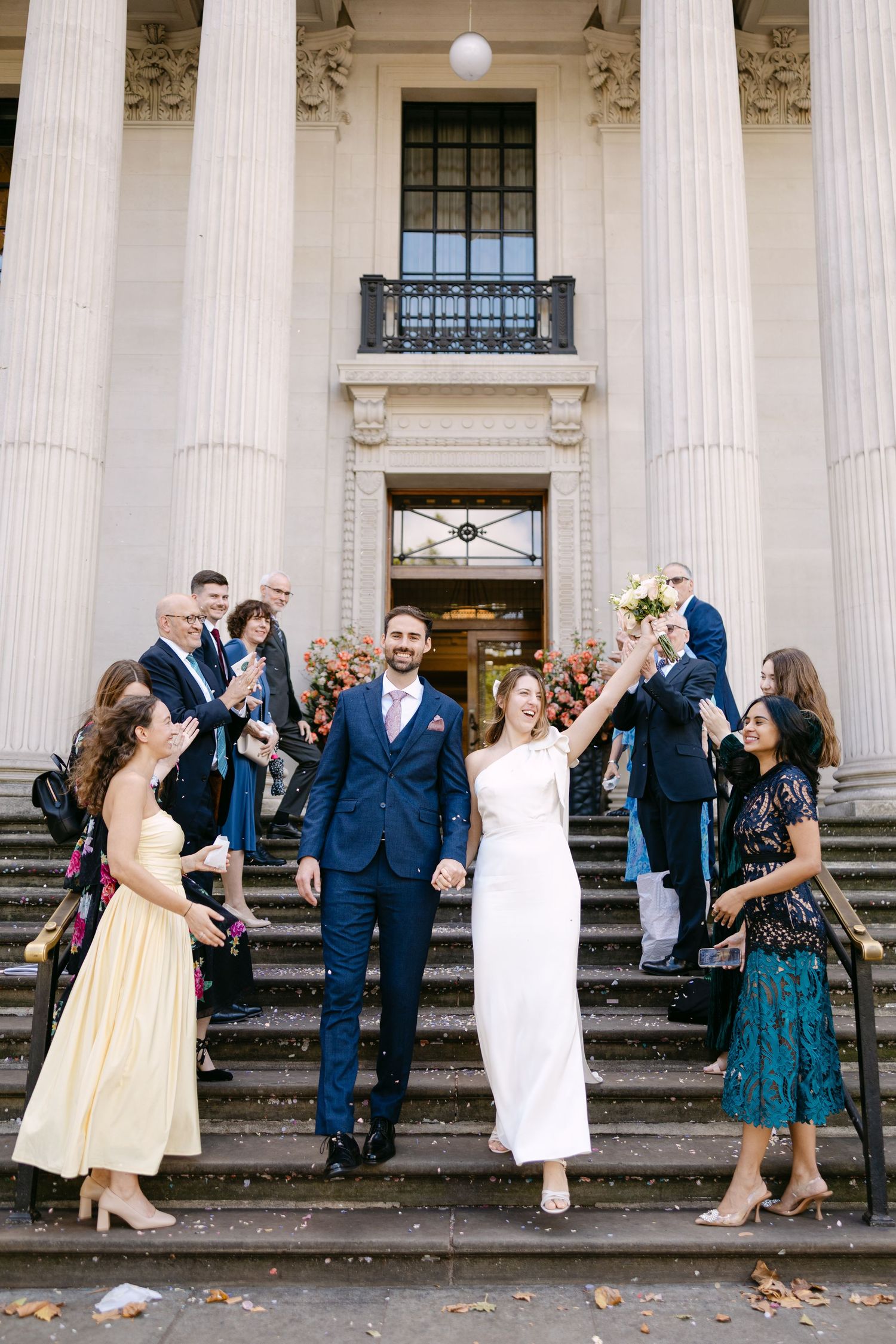Wedding party celebrates on historic building steps with white columns, creating a joyful scene with dancing and laughter.