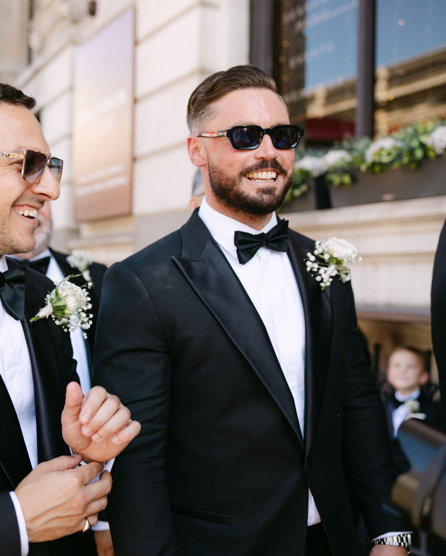 Groomsmen in black tuxedos and sunglasses smile and laugh together during outdoor wedding photos.