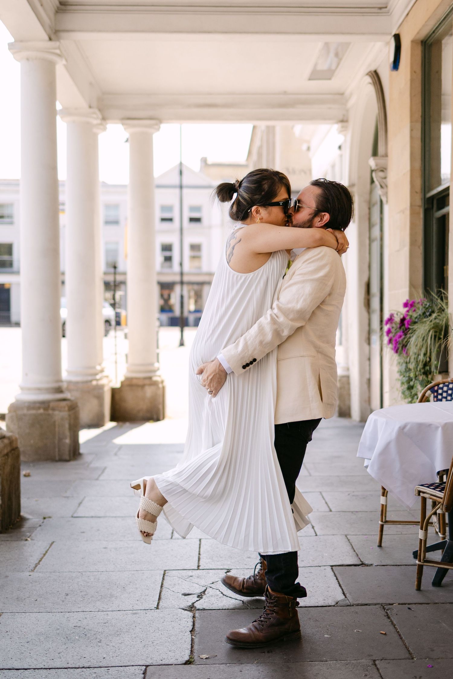 Couple sharing a romantic kiss under white columns, woman in pleated white dress lifted by man in beige blazer.