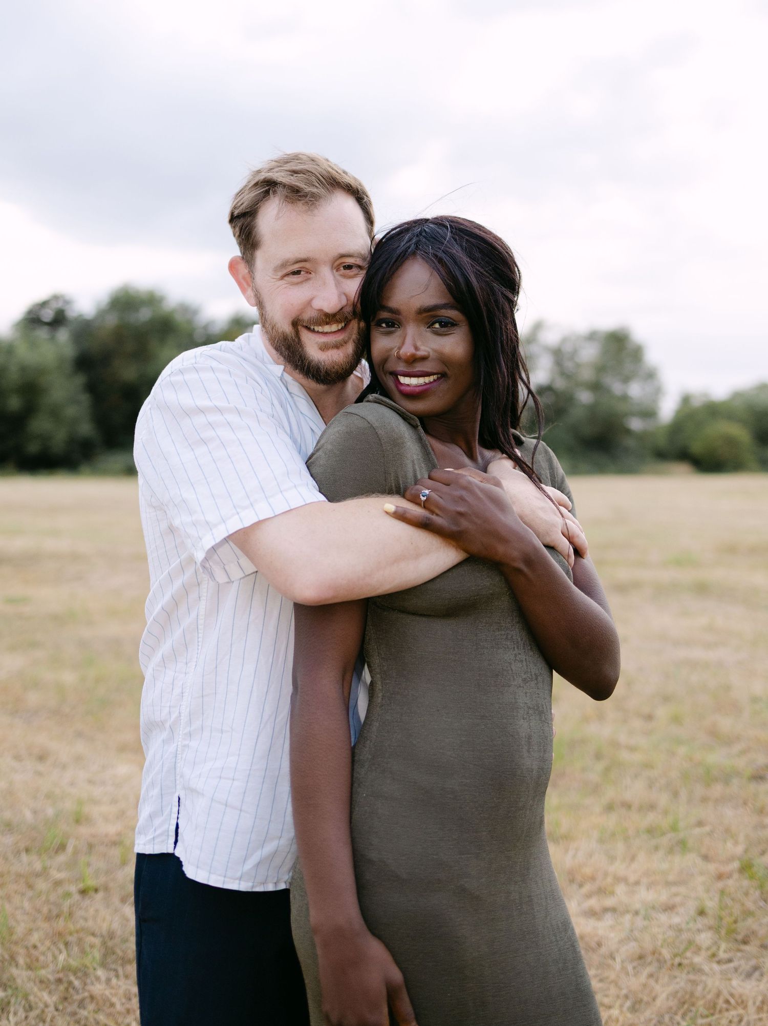 A couple embraces and laughs together in a sun-lit grassy field on a warm summer day.