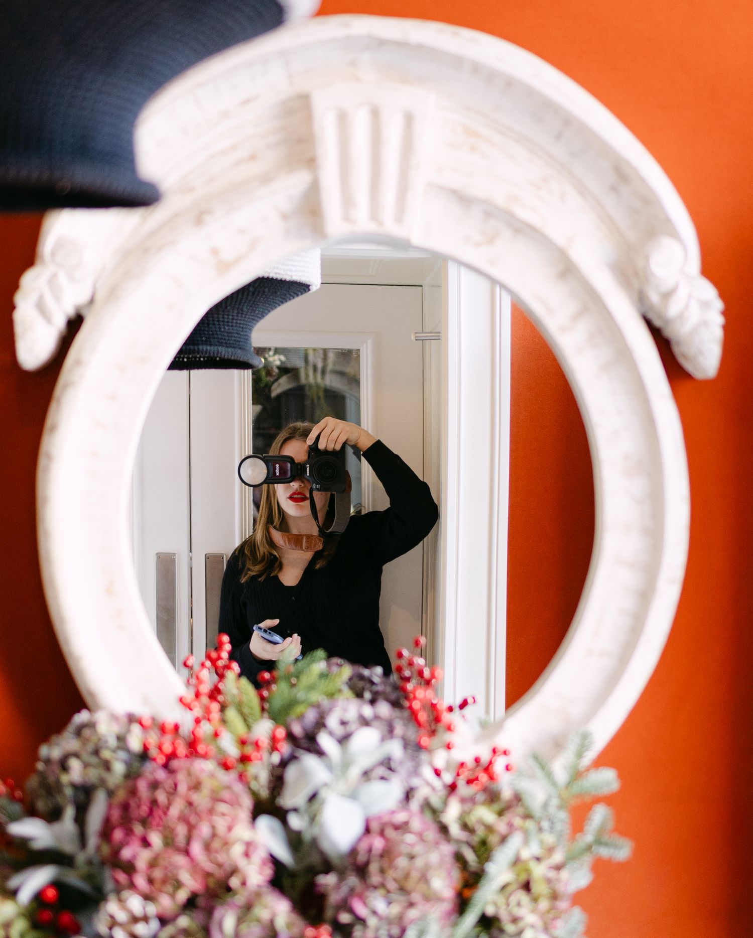 Photographer takes a selfie in an ornate white mirror frame with pink floral arrangements against a bright orange wall.