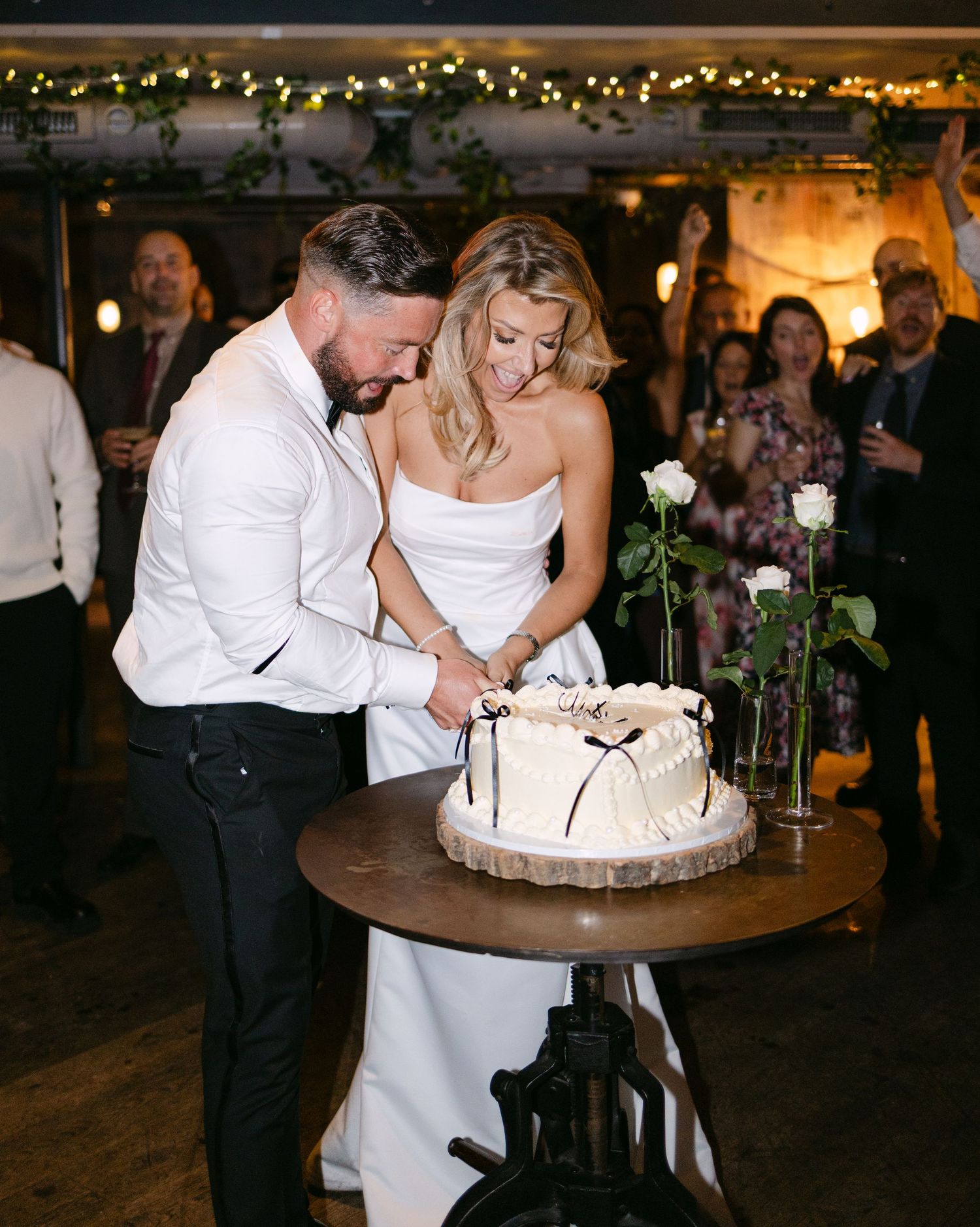 A couple cuts their elegant white wedding cake together at a rustic reception venue with string lights overhead.