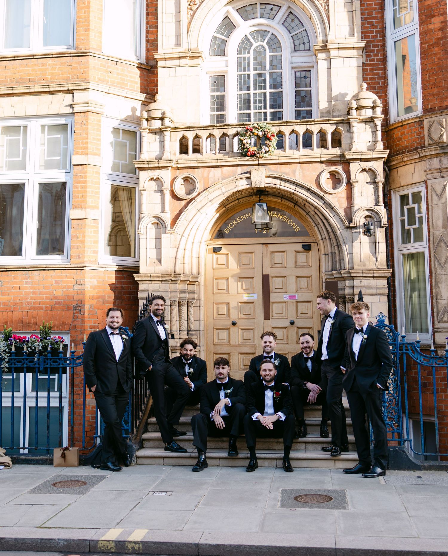 Groomsmen in tuxedos pose on steps of ornate Victorian building entrance.