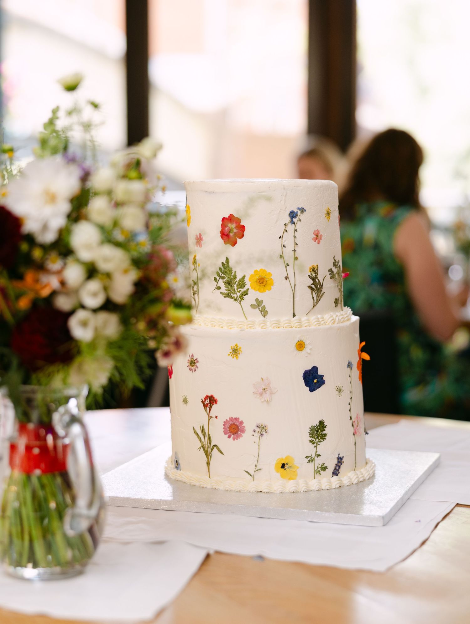 An elegant white wedding cake decorated with pressed wildflowers sits on display with floral arrangements.