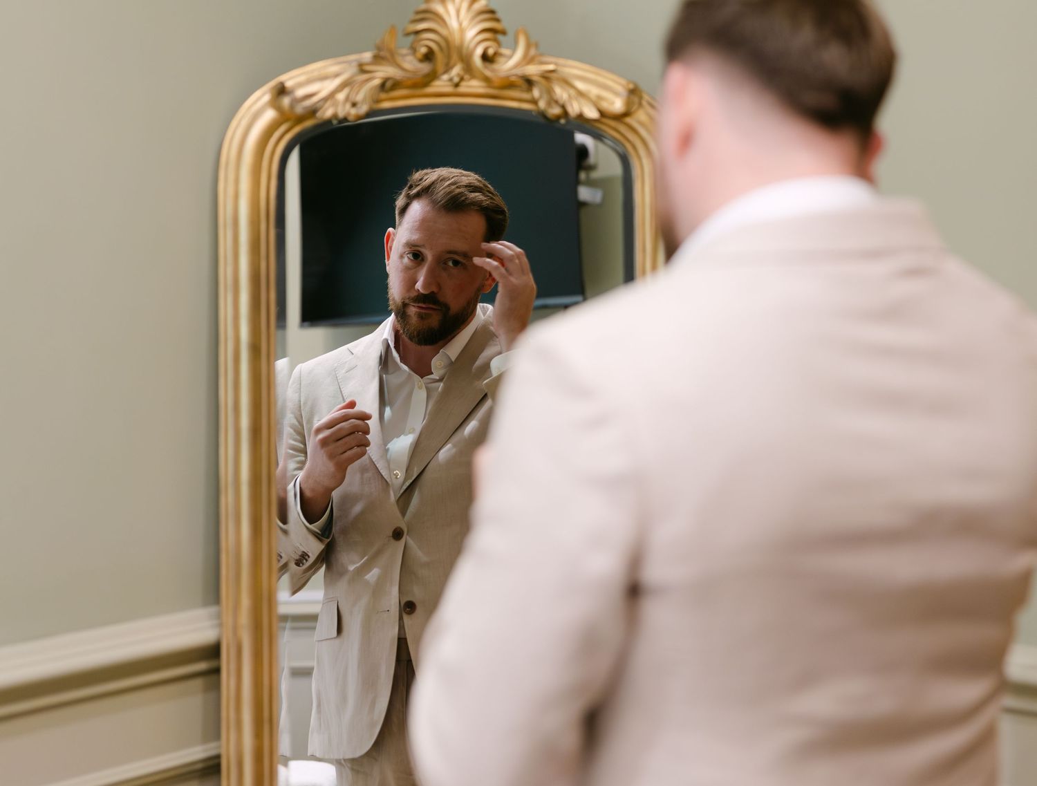 Person in light suit adjusting their appearance in ornate gold mirror.