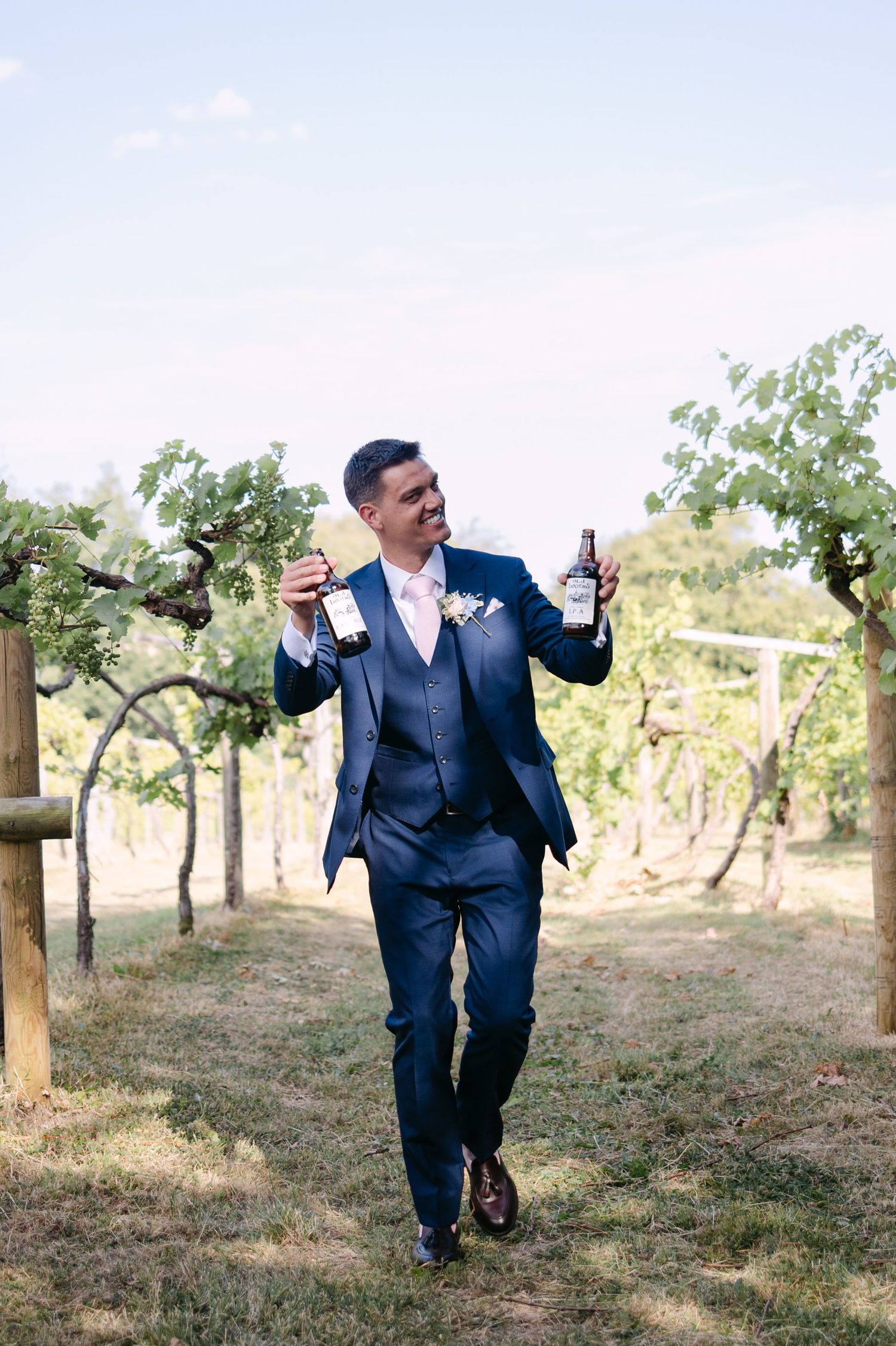 Groom in navy suit holds two wine bottles while walking through a sunny vineyard.
