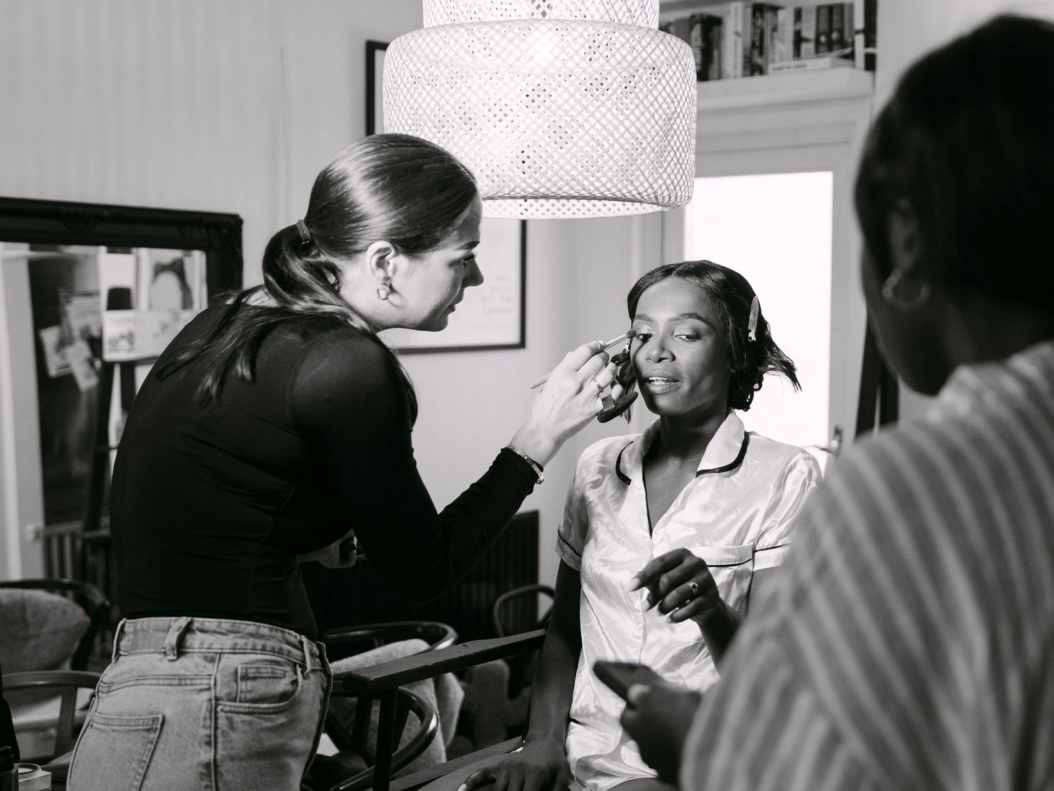 Black and white photo of a makeup artist applying makeup to a seated woman in a bright, stylish room.