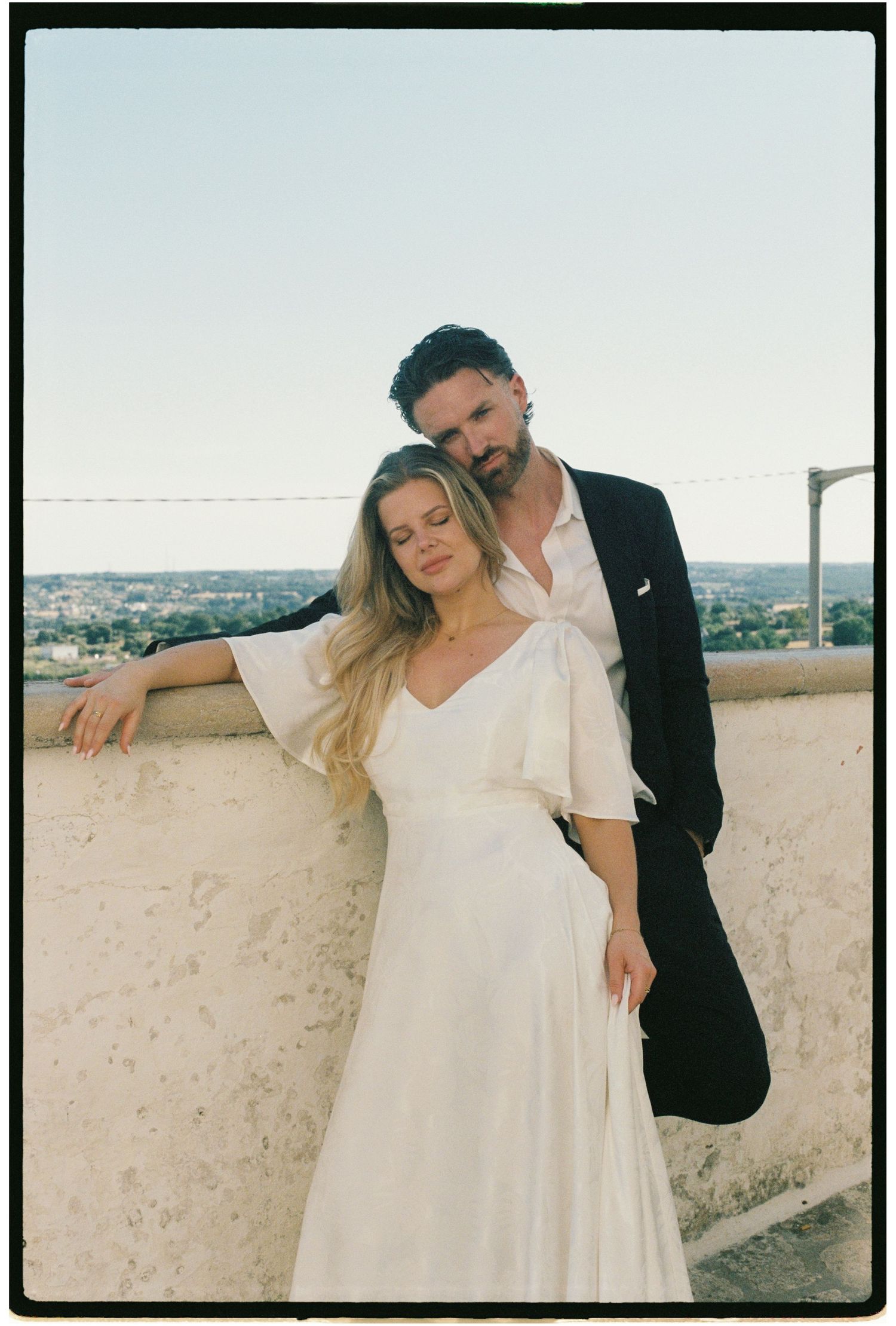 Couple embracing on a rooftop terrace overlooking a sunny cityscape, woman in white flutter dress.